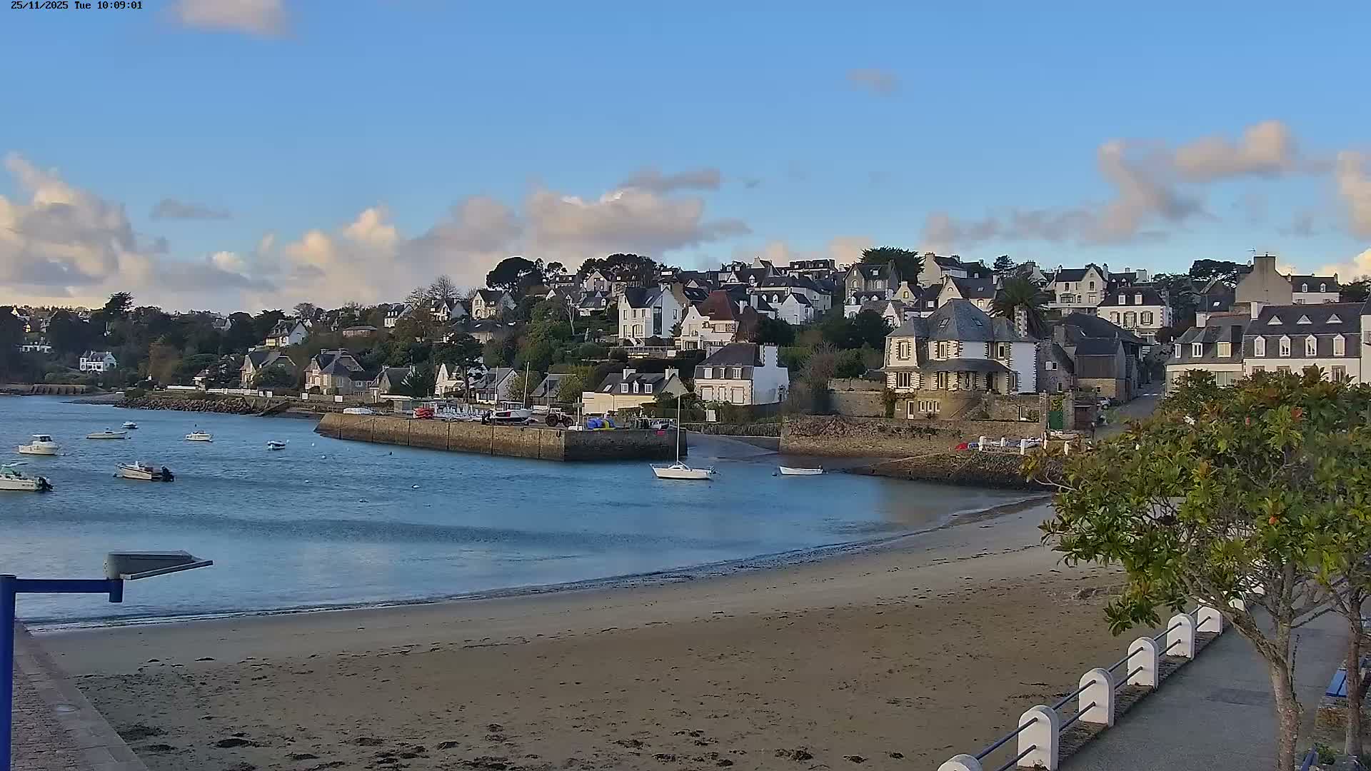 A picturesque coastal town featuring numerous houses nestled on a hillside overlooks a calm bay with several boats and a sandy beach under a partly cloudy blue sky.
