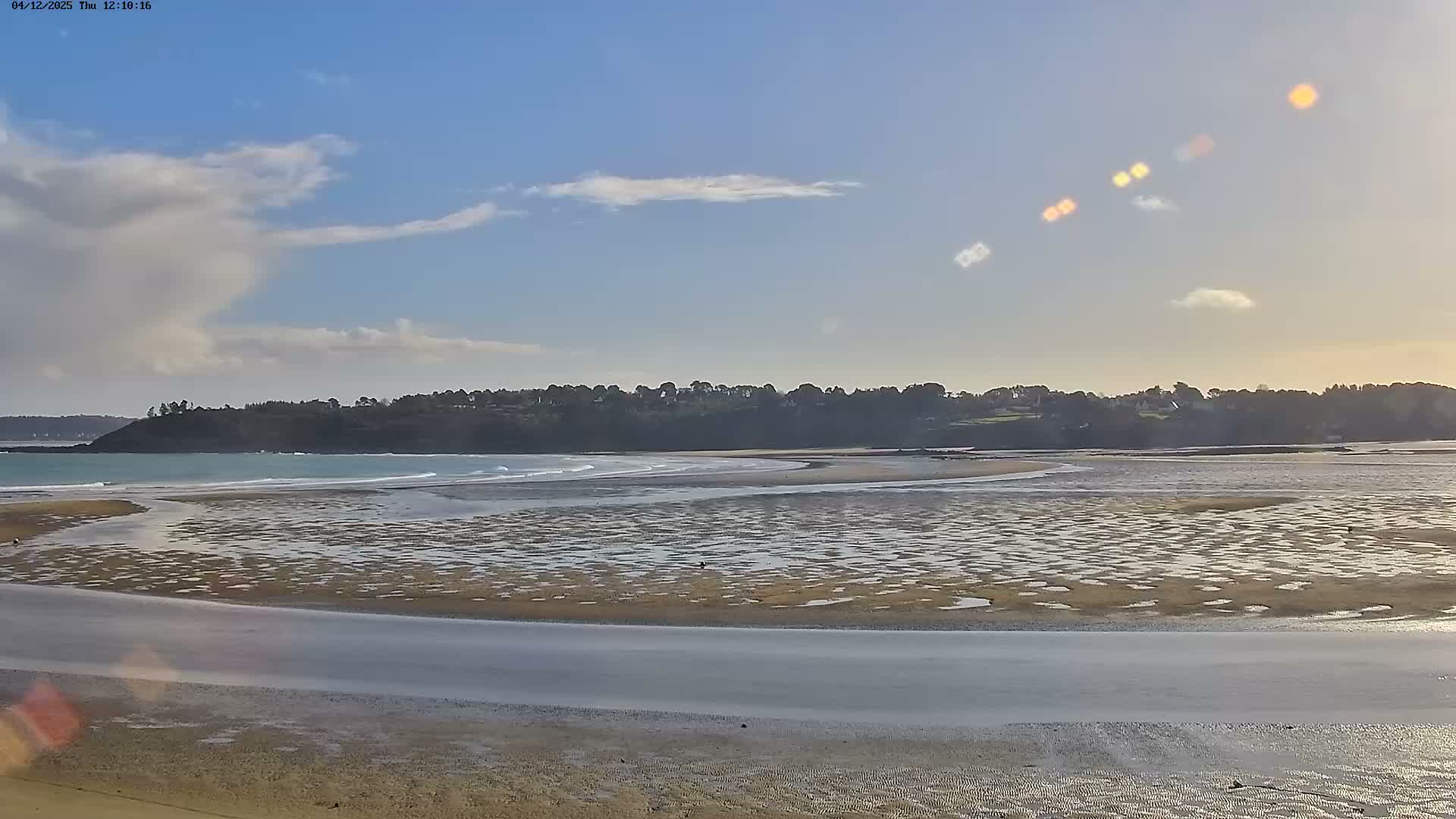 A vast sandy beach at low tide extends into calm blue waters under a bright, partly cloudy sky, with a tree-covered coastline in the distance.