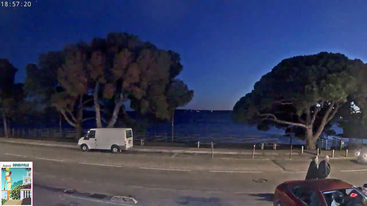 A white van is parked on the side of a road next to a body of water under a twilight sky, with several large trees and two people walking near a red car.