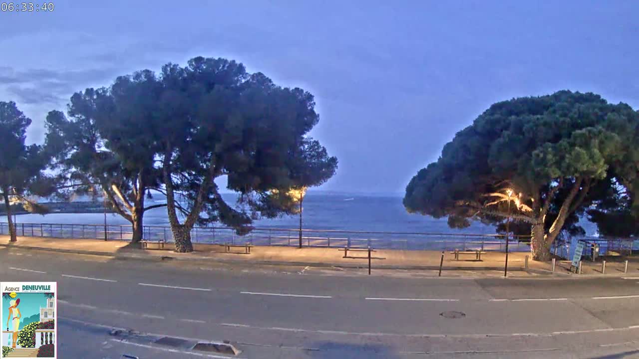 A calm, dark blue body of water is visible beyond a paved walkway lined with trees and benches under a twilight sky.