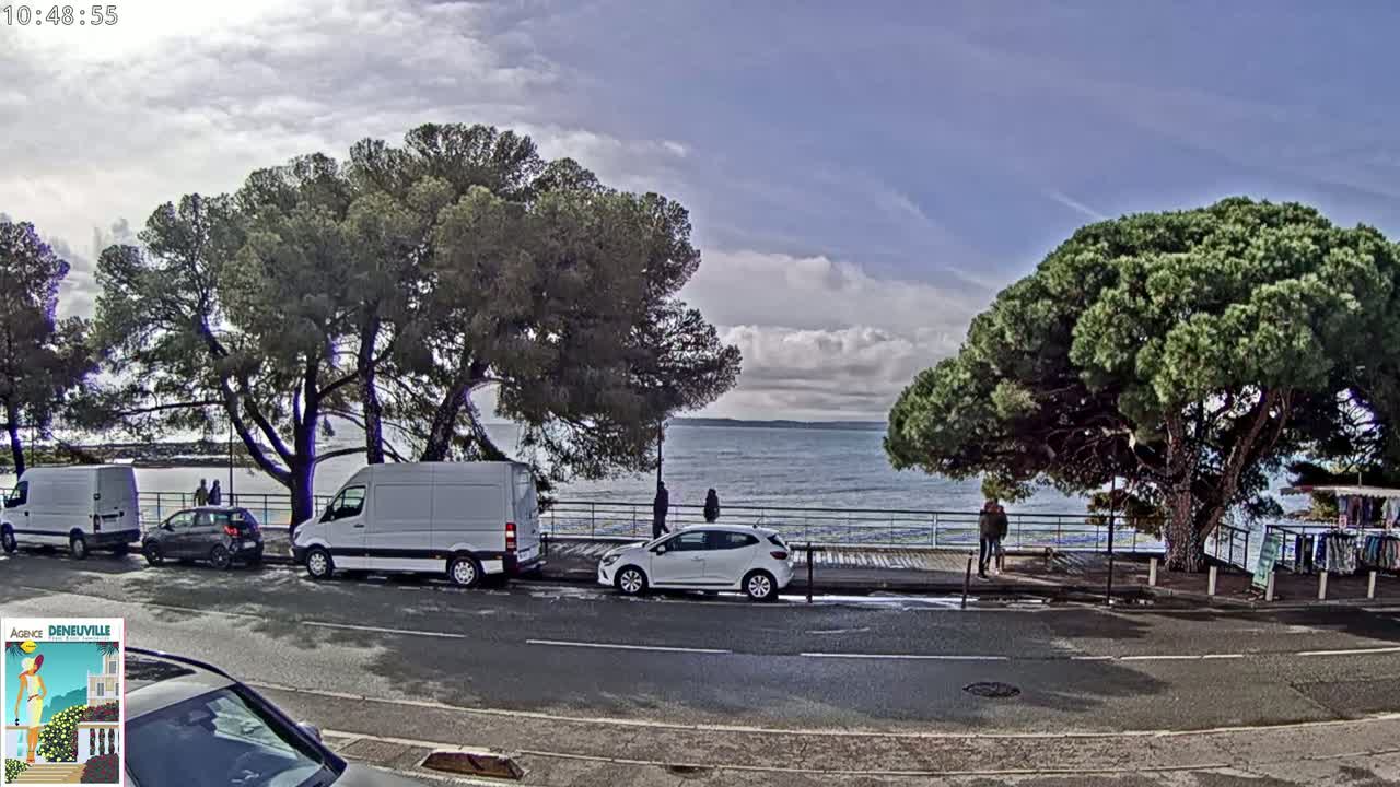 A partly cloudy day reveals a seaside scene with several parked cars and vans near a boardwalk lined with trees, overlooking a calm body of water.