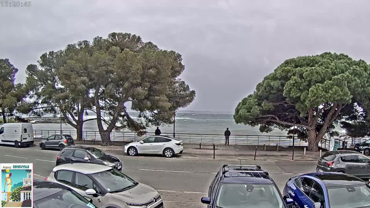 A parking lot with several cars is situated next to a shoreline with some choppy waves under an overcast sky, and two people are standing by the railing overlooking the water.