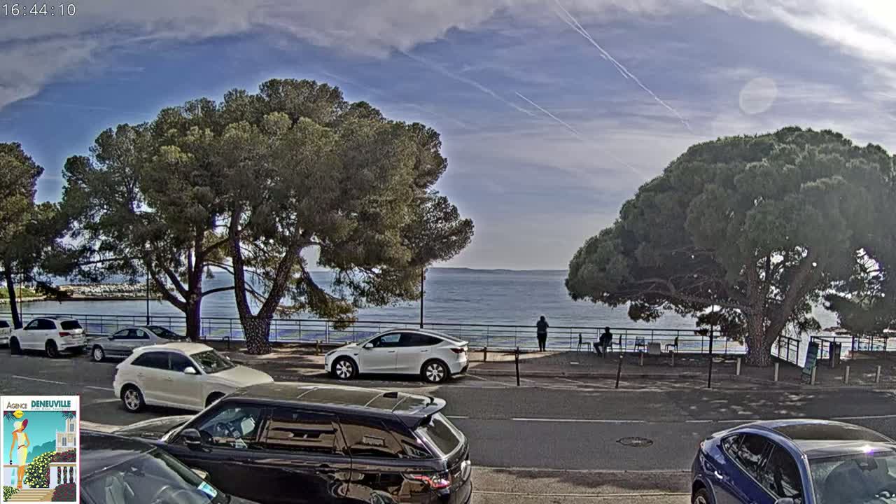 A partly sunny day overlooks a calm body of water, where several cars are parked along a road lined with trees and a few people are standing by a railing.