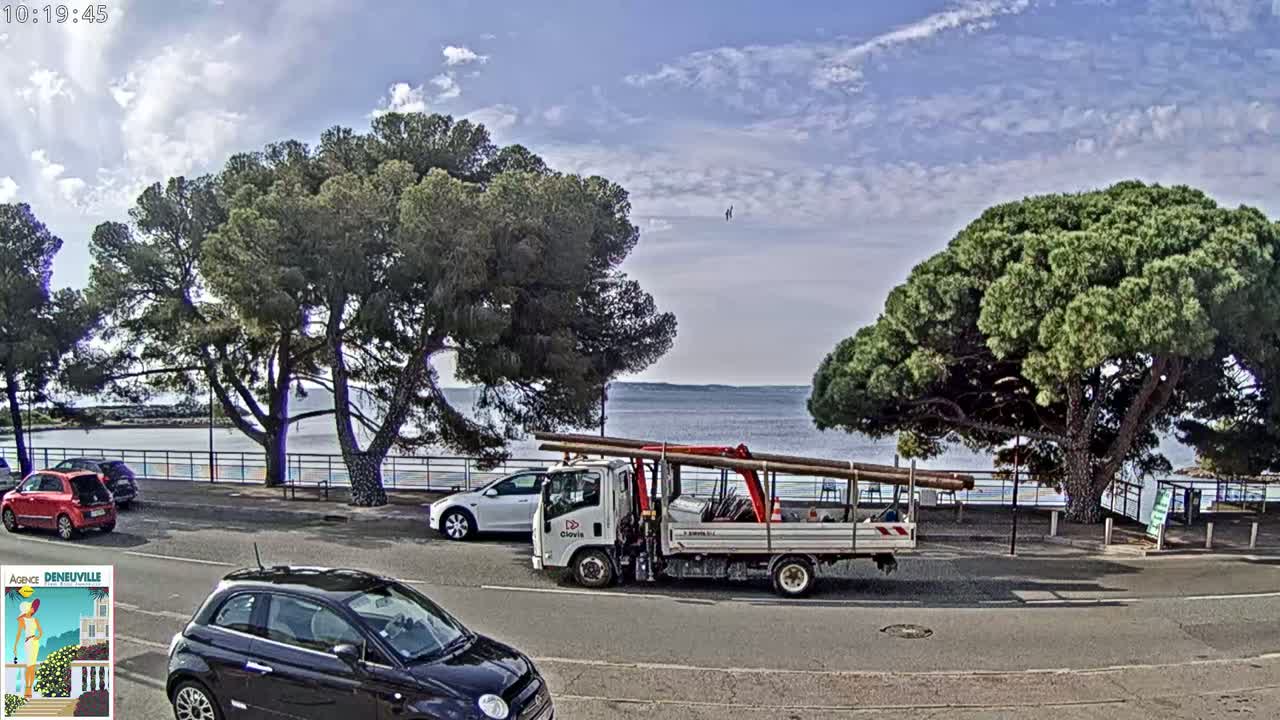 A partly sunny day shows a seaside parking lot with several cars, a flatbed truck carrying long beams, and large pine trees overlooking a calm body of water.