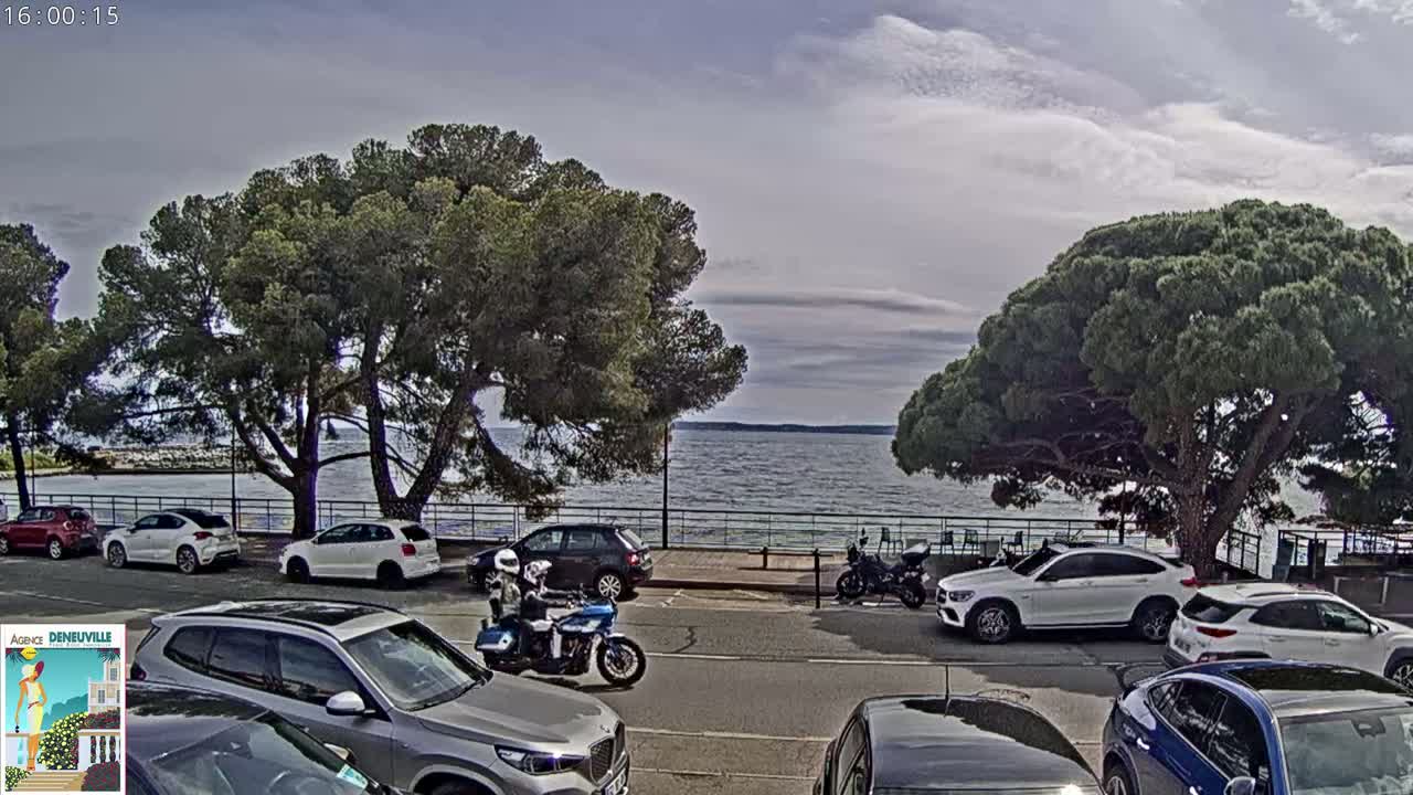 Under a partly cloudy sky, cars are parked along a road next to a calm lake, with several large pine trees and two motorcyclists visible.