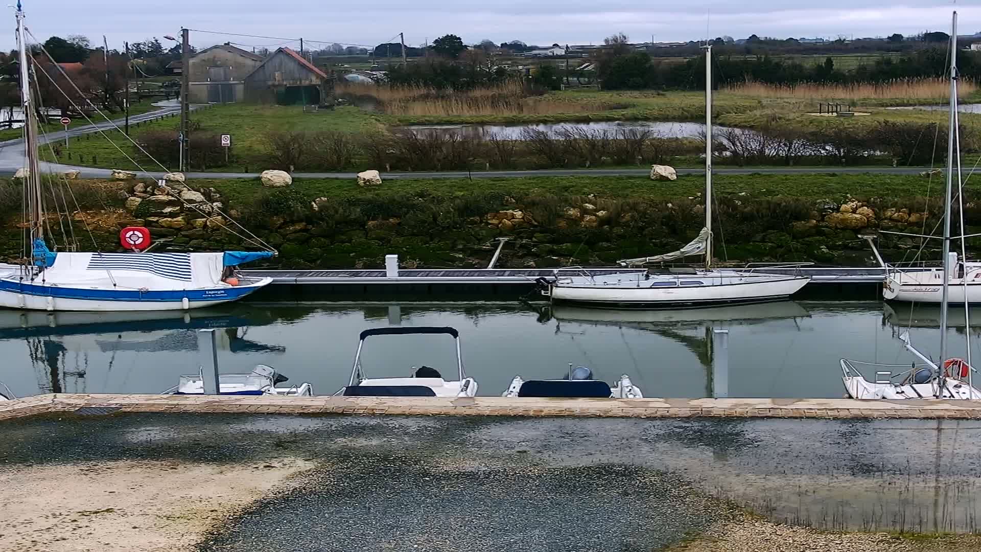 Several sailboats and motorboats are moored at a dock along a calm waterway under an overcast sky, with green fields and a building visible in the background.