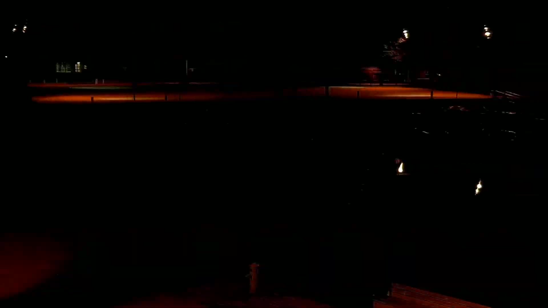 A dark, nighttime scene shows an illuminated flat, open area with a few distant lights and a building faintly visible in the background.