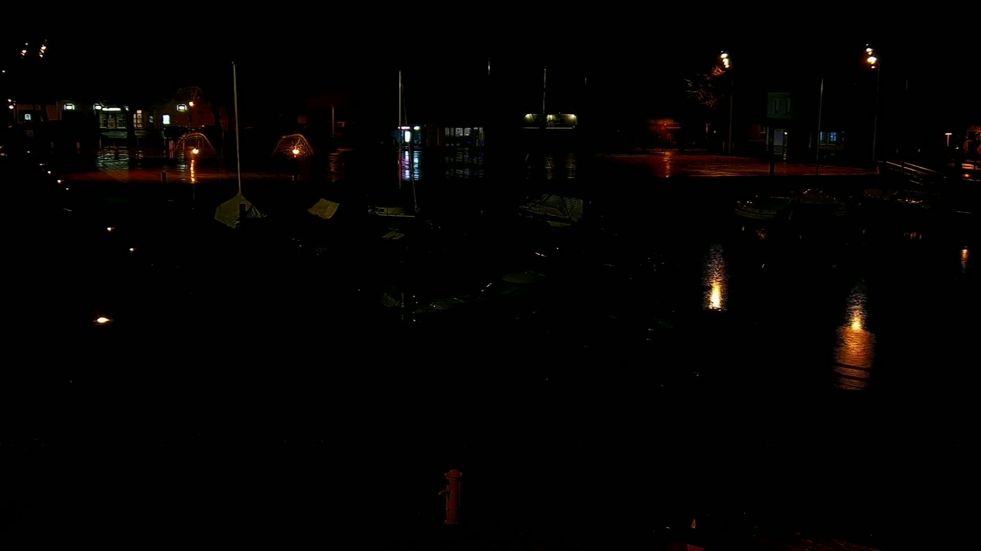 Several boats are moored in a dark, wet harbor at night, illuminated by streetlights and reflections on the water.