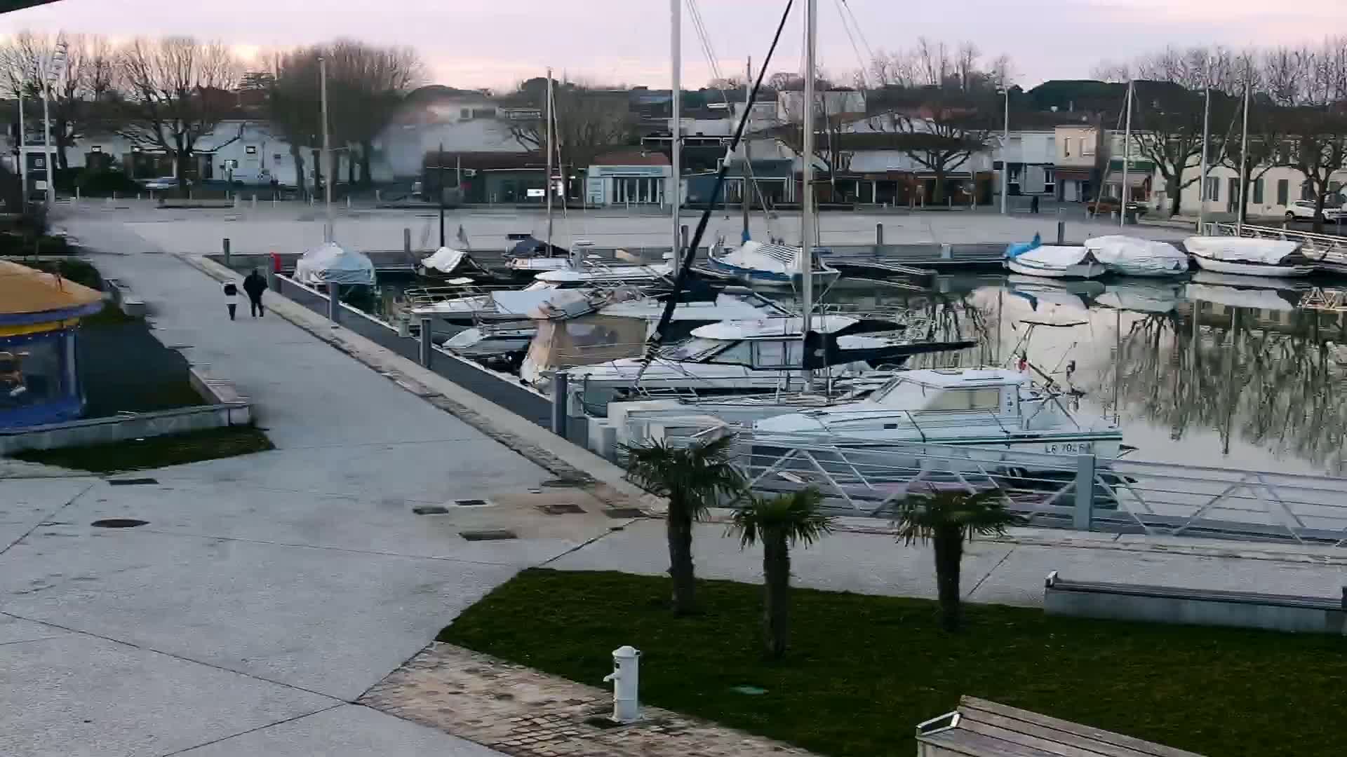 A calm marina filled with boats under a twilight sky, with two people walking along a paved walkway.