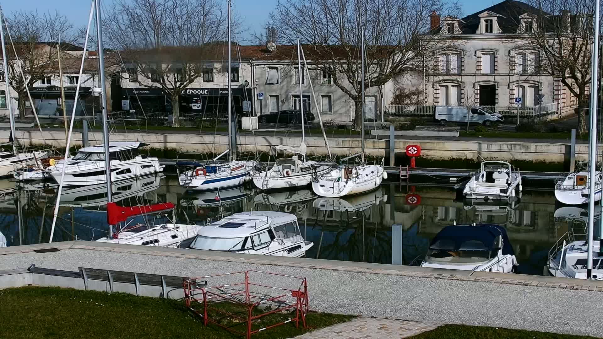 Several sailboats and motorboats are docked in a calm harbor on a sunny day, with buildings lining the waterfront.