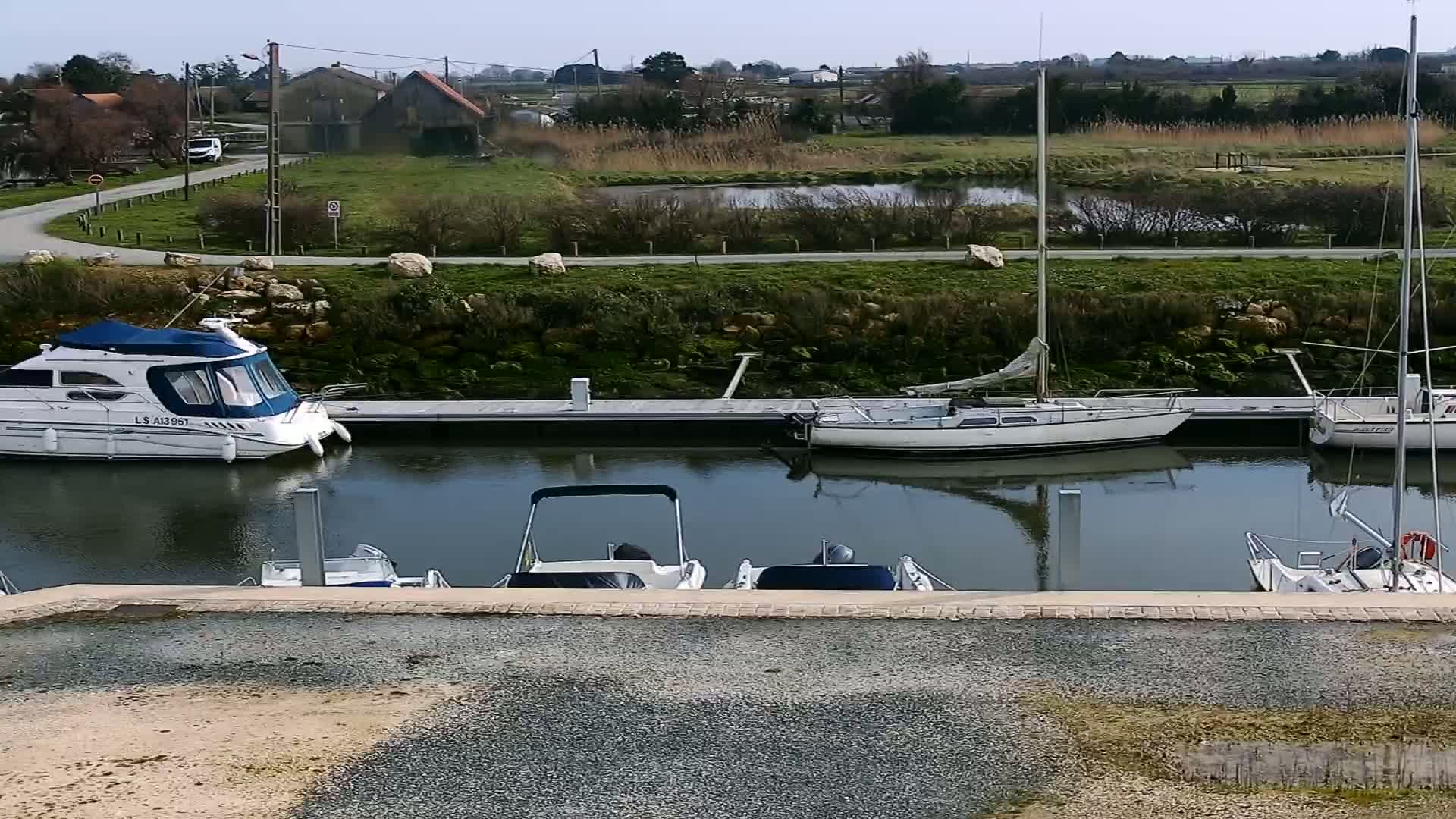 Several boats are moored at a marina on a calm waterway, under a clear sunny sky, with a grassy area and buildings visible in the background.
