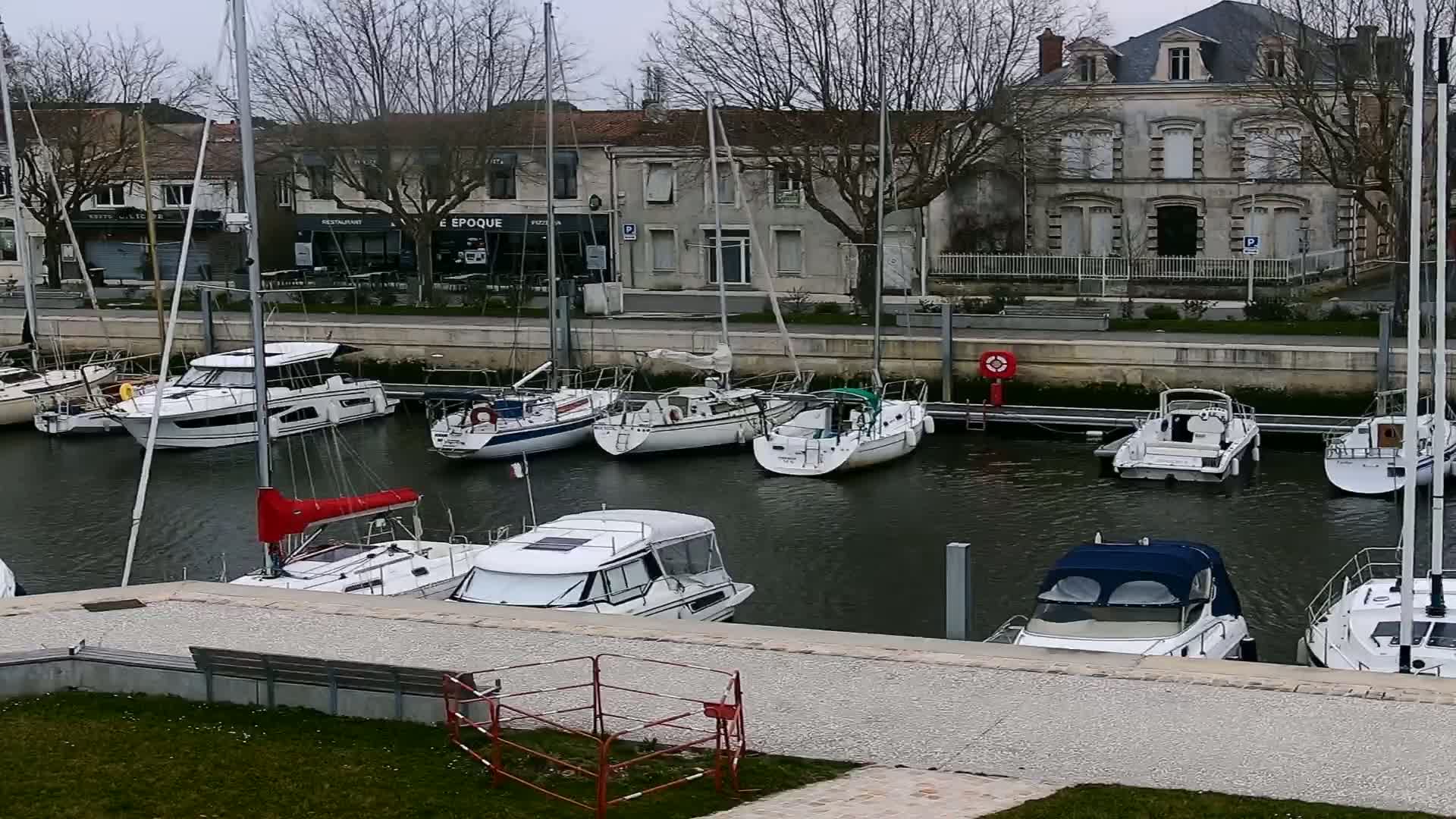 A marina filled with sailboats and motorboats is docked along a waterfront lined with buildings under an overcast sky.