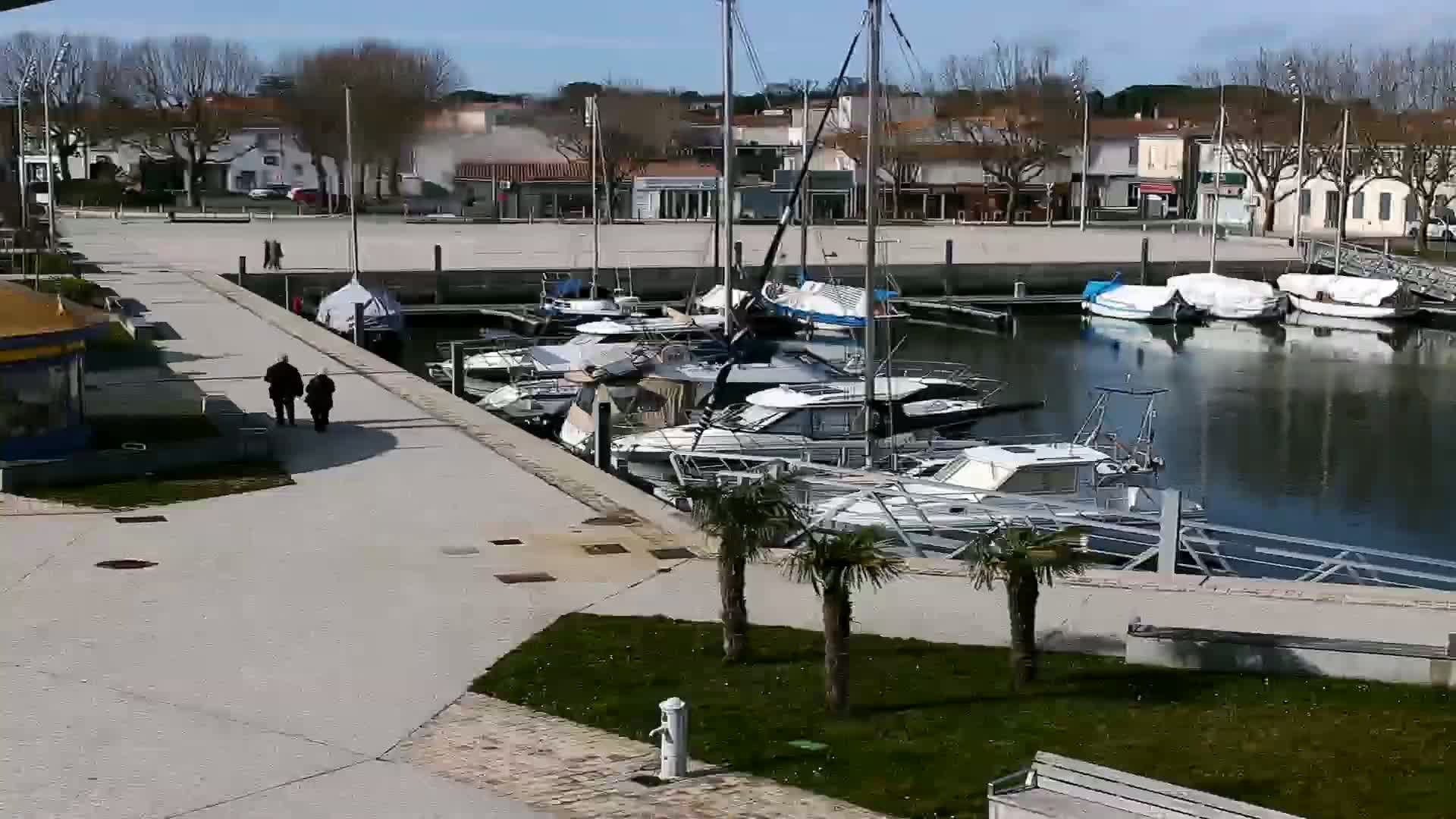 A marina filled with boats is seen on a sunny day, with two people walking along a paved walkway beside it.