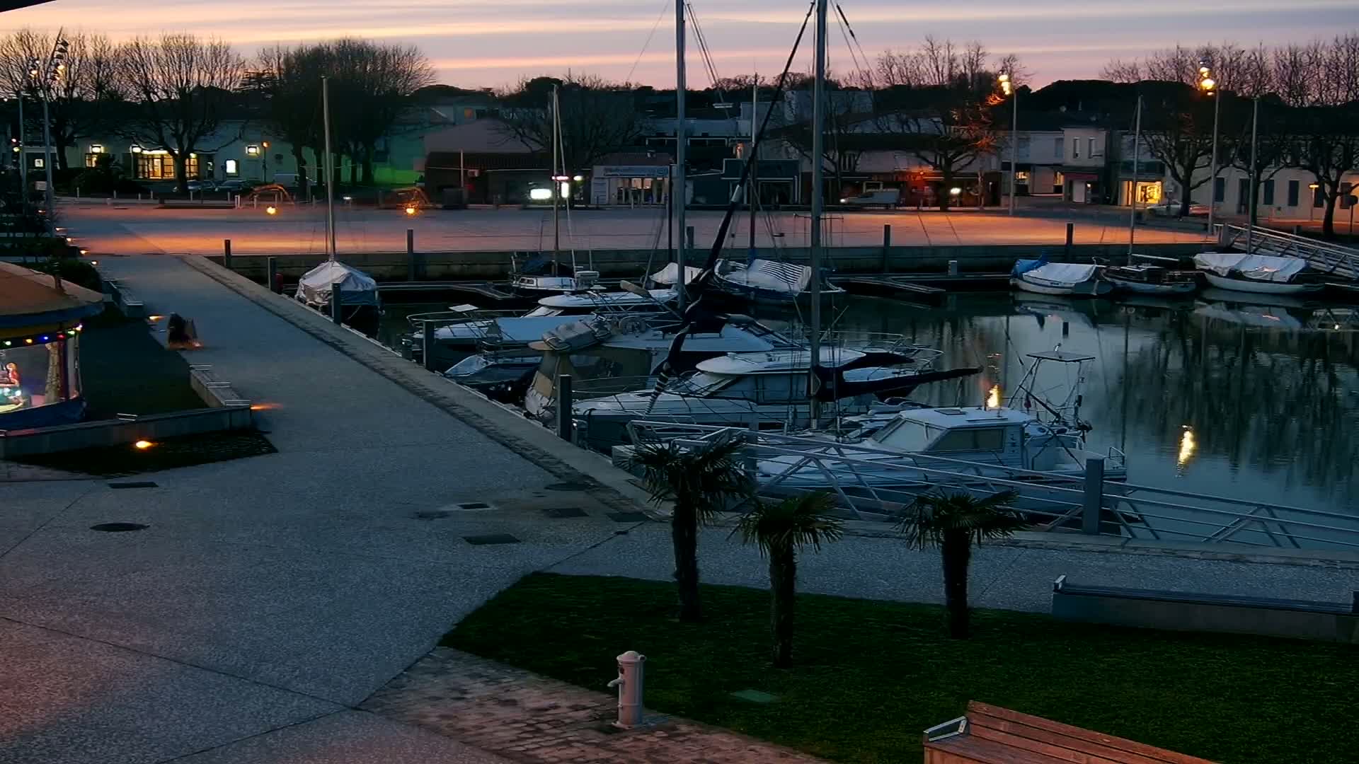 A marina at twilight, filled with numerous boats, is situated beside a paved walkway and a small park next to a town.