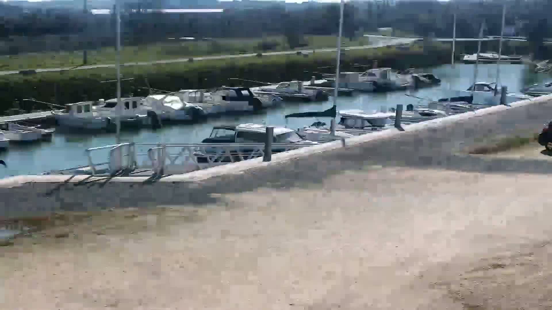 Numerous small boats are moored in a calm marina next to a dirt road and a low concrete wall.