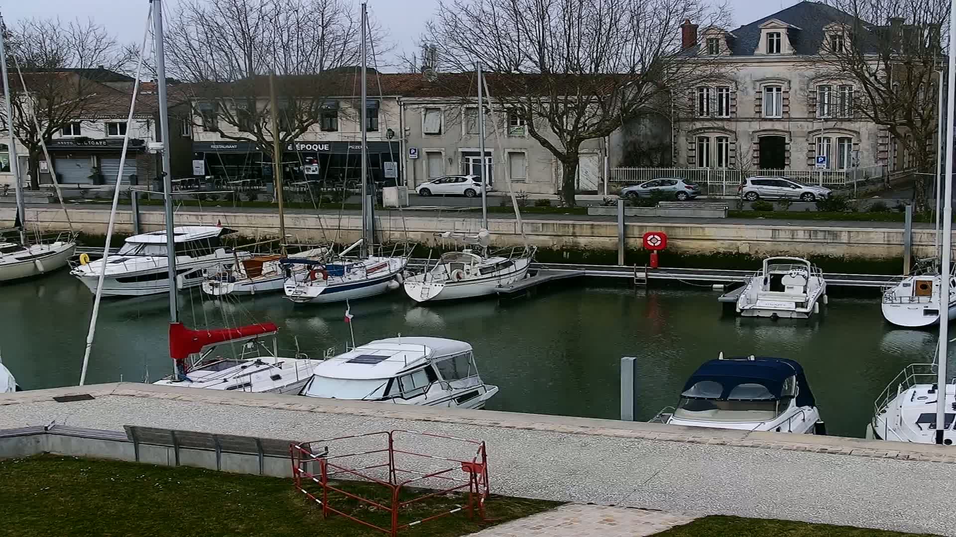 A marina filled with sailboats and motorboats sits in front of a row of buildings on an overcast day.