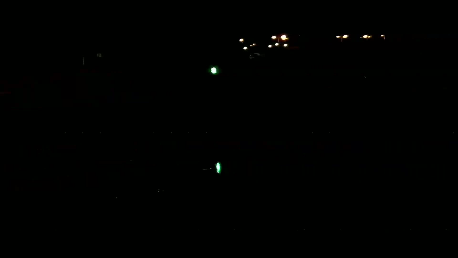 A dark nighttime scene shows a few distant lights and a faint green light reflected on a dark body of water.