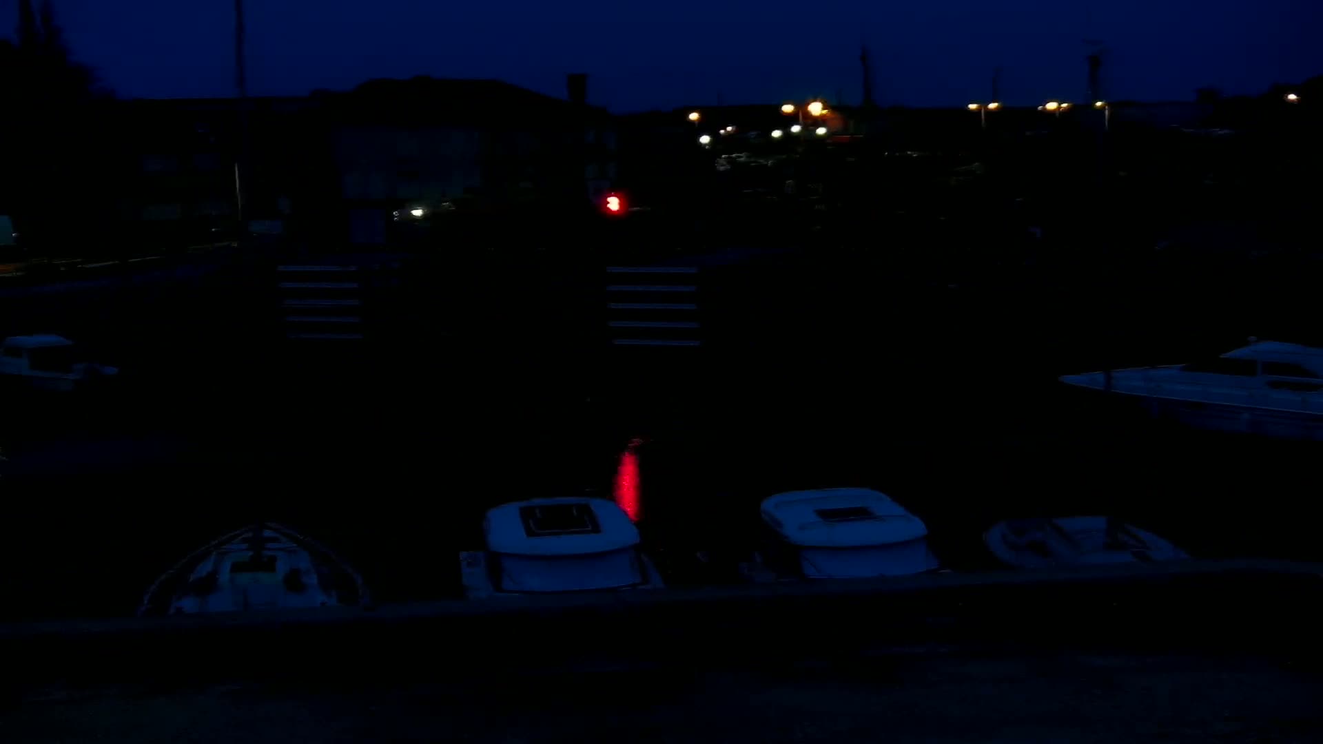 Several small boats are moored in a dark marina at night under a dark blue sky, illuminated by a few distant lights and a single red light on the water.