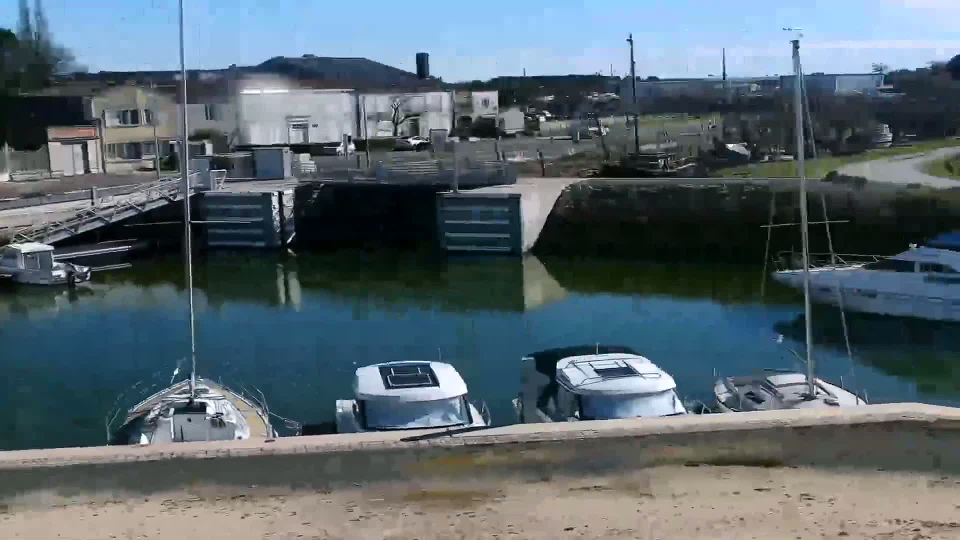 Several boats are moored in a calm, green-tinged waterway next to a small town on a sunny day.