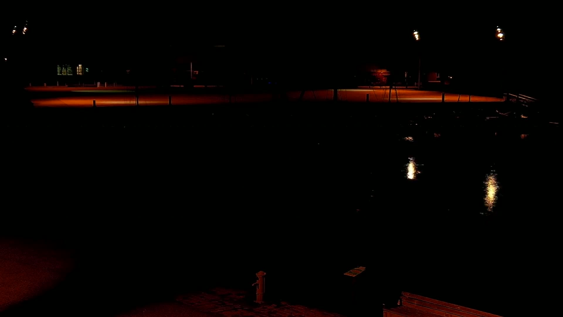 An empty, dimly lit dock area at night is seen next to a body of dark water reflecting the lights.