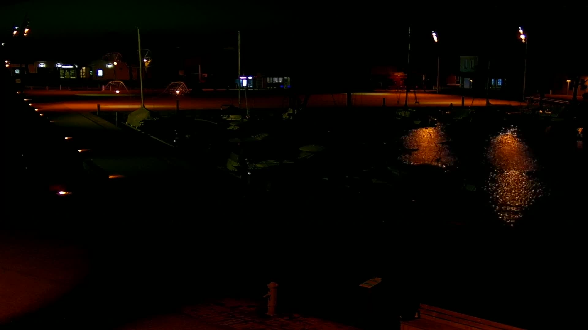 At night, a dark harbor is illuminated by lights reflecting on the water, with several boats moored and a paved area visible in the background.