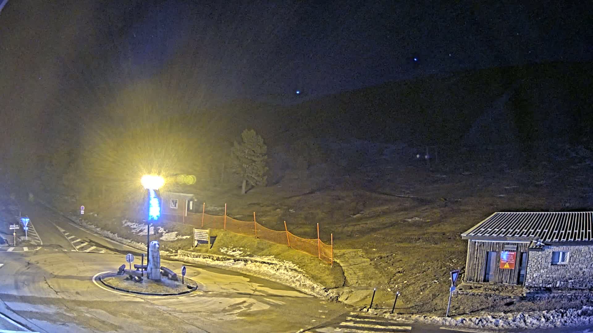 A nighttime scene shows a roundabout with a statue at its center, a small building, and an orange safety fence bordering a bare, dark hill under a mostly clear, dark sky with a few visible stars.