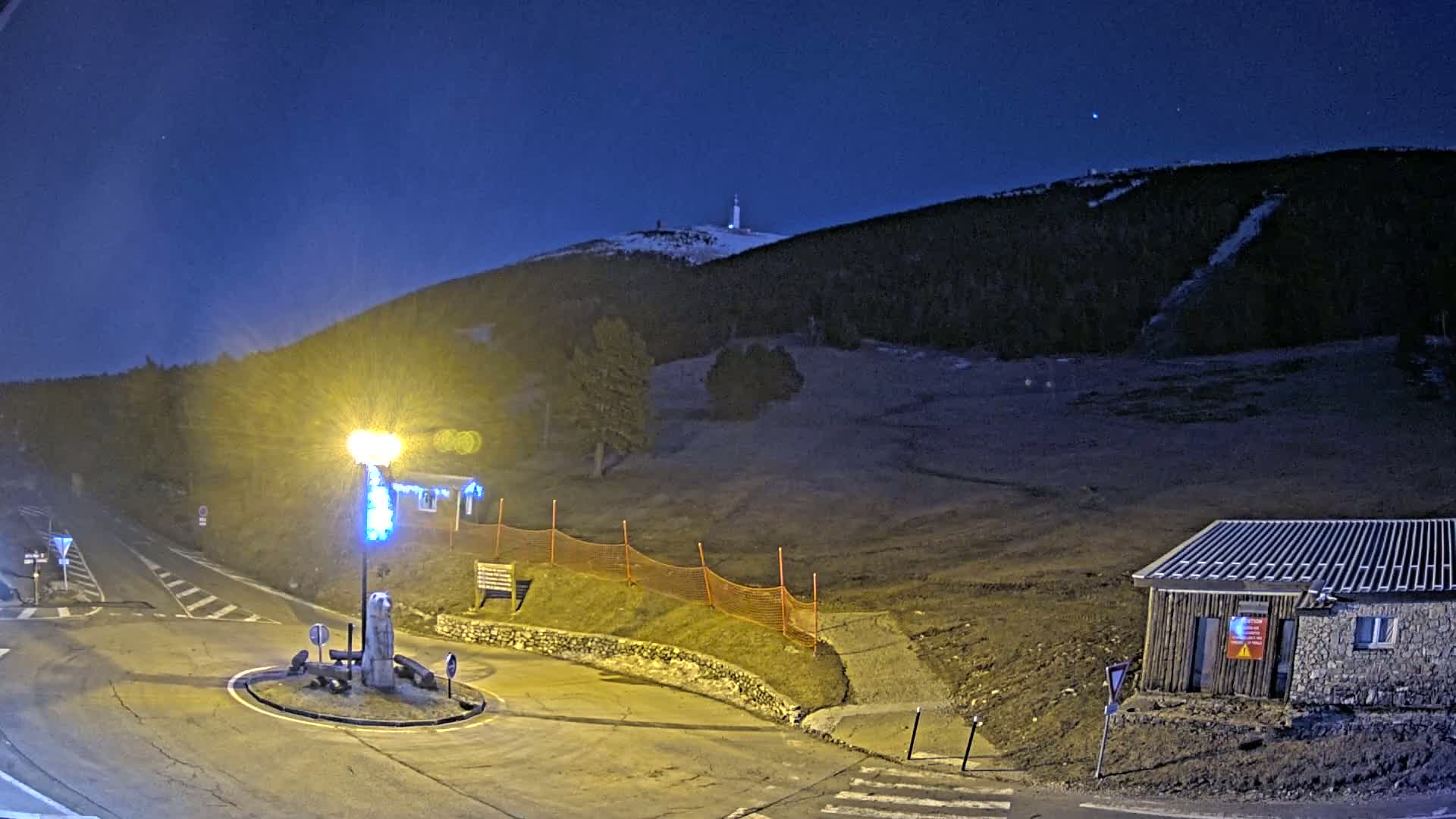 A nighttime view of a snow-dusted hill with a tower on top, a small building, and a roundabout illuminated by a streetlight.