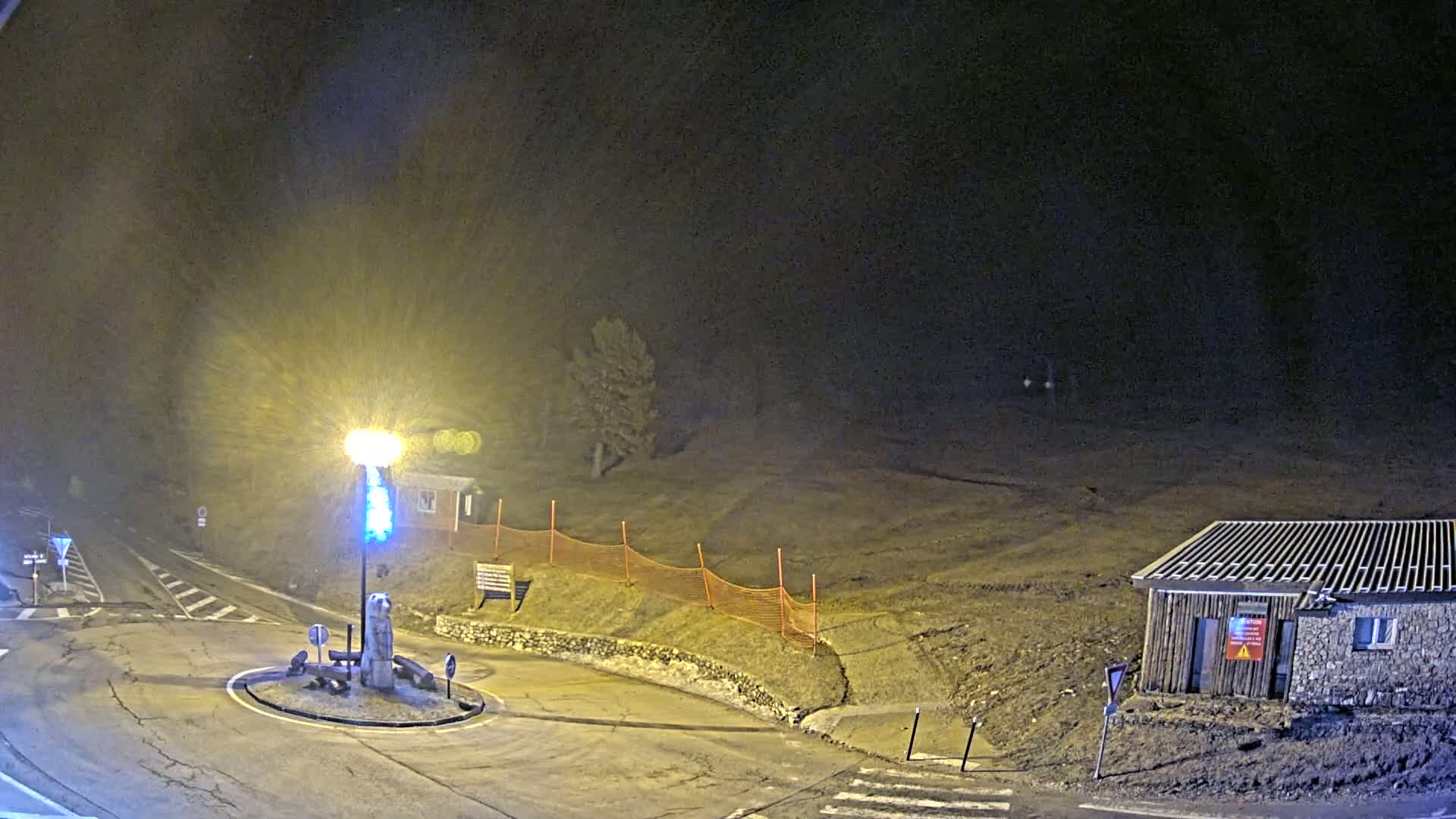A nighttime scene shows a roundabout with a lit lamppost, a small building, and an orange safety net, under snowy conditions.