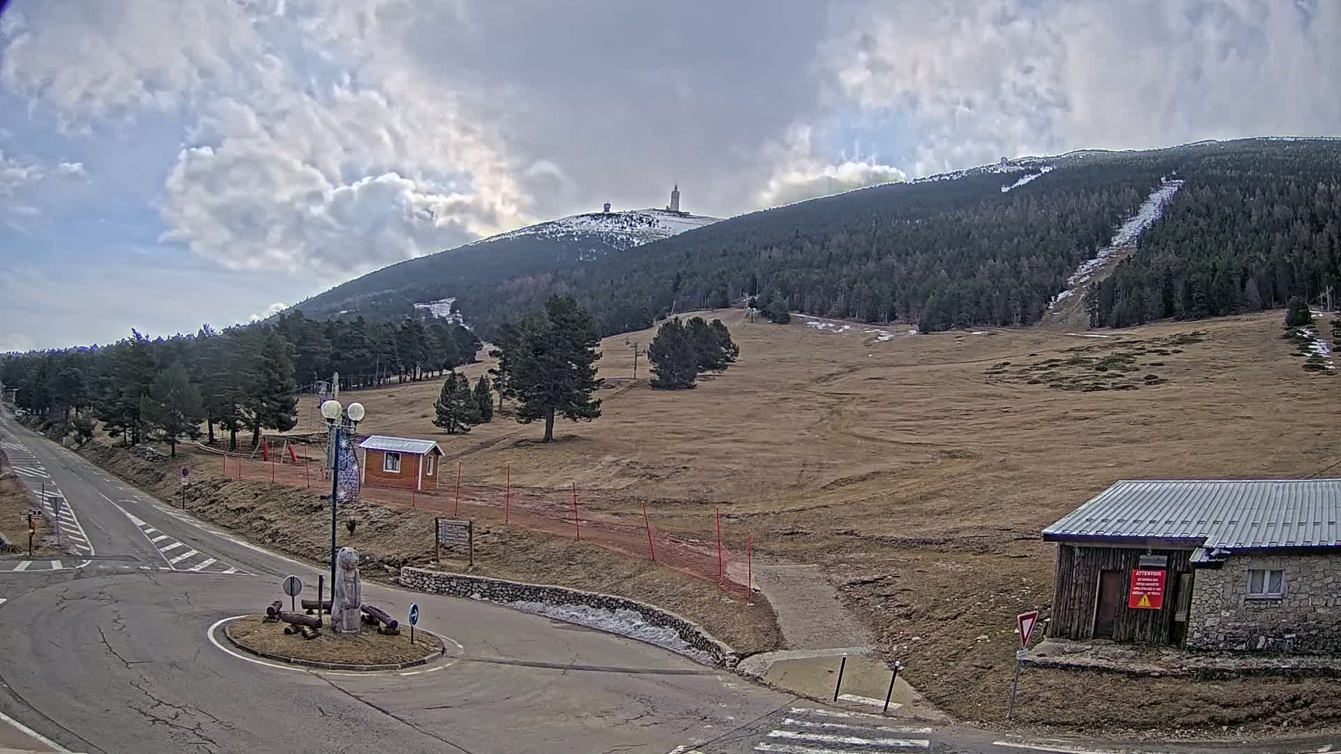 A mountaintop with a tall structure is visible in the distance under a partly cloudy sky, overlooking a sparsely vegetated landscape with a road, small buildings, and a roundabout.