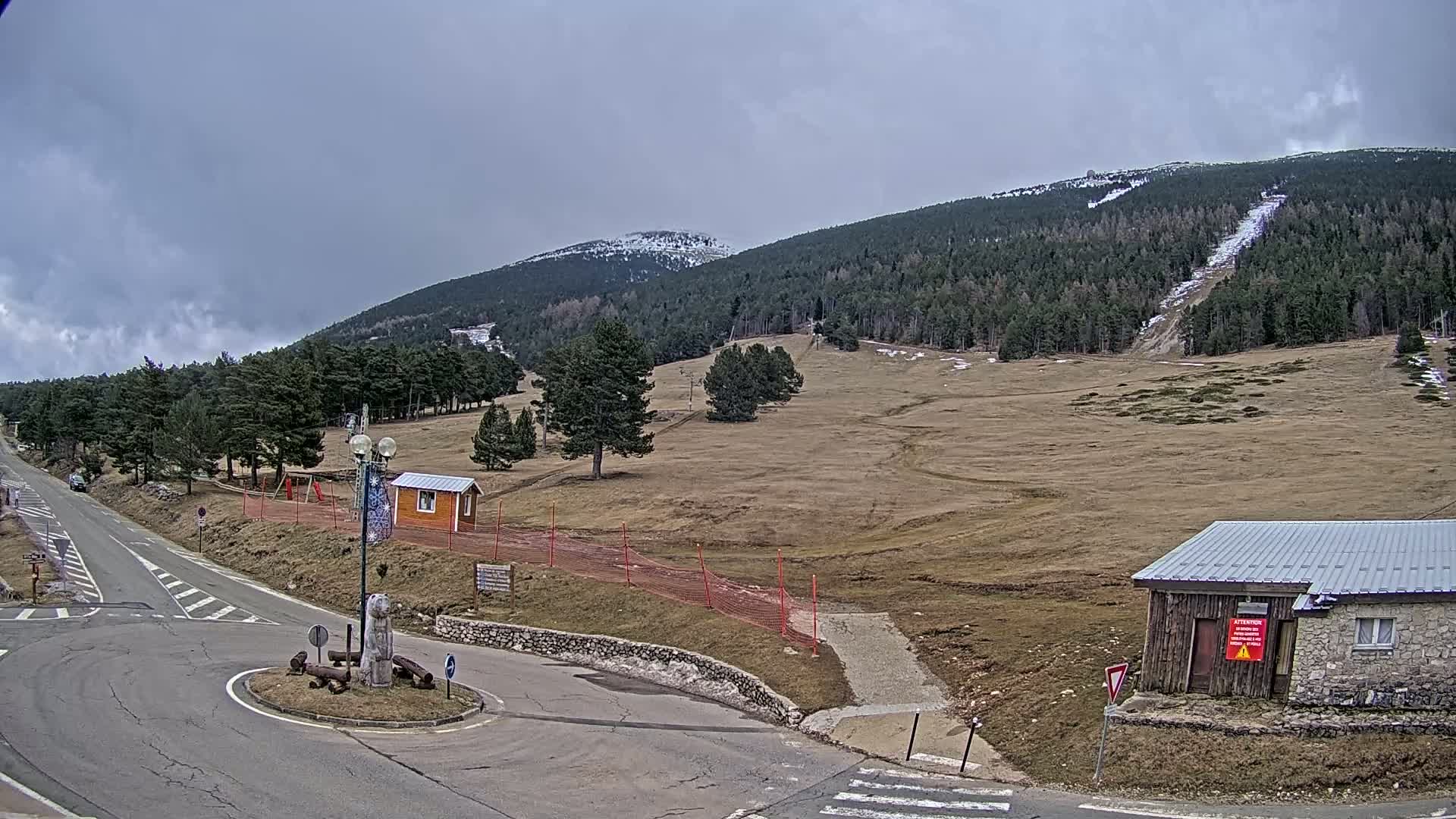 A mountaintop scene under a cloudy sky shows a road curving through a sparsely vegetated area with a few buildings, some trees, and a ski lift in the distance.