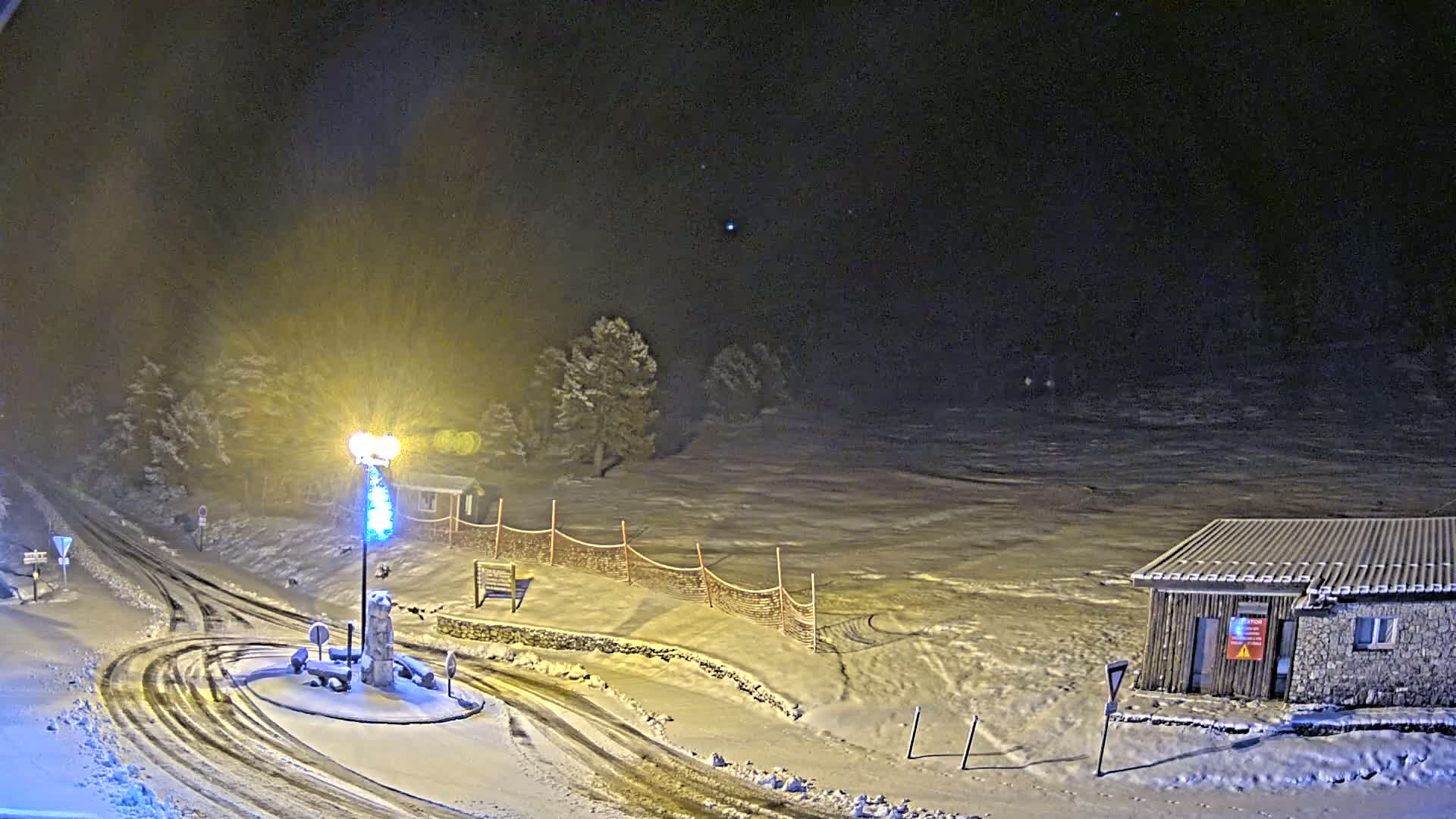 A snow-covered landscape at night, illuminated by a lamppost, shows a small building, a snow-covered roundabout, and a section of what appears to be a ski slope.