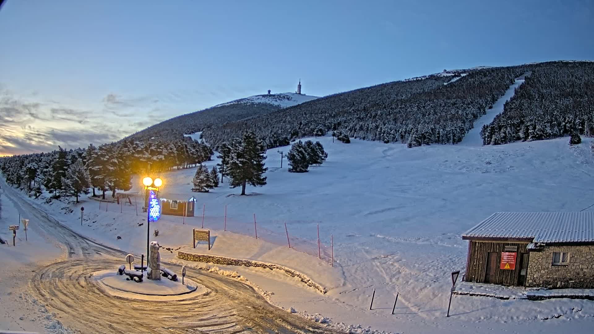 A snow-covered ski resort at dusk, with a lit lamppost in the foreground and a tall tower visible on the distant mountain.