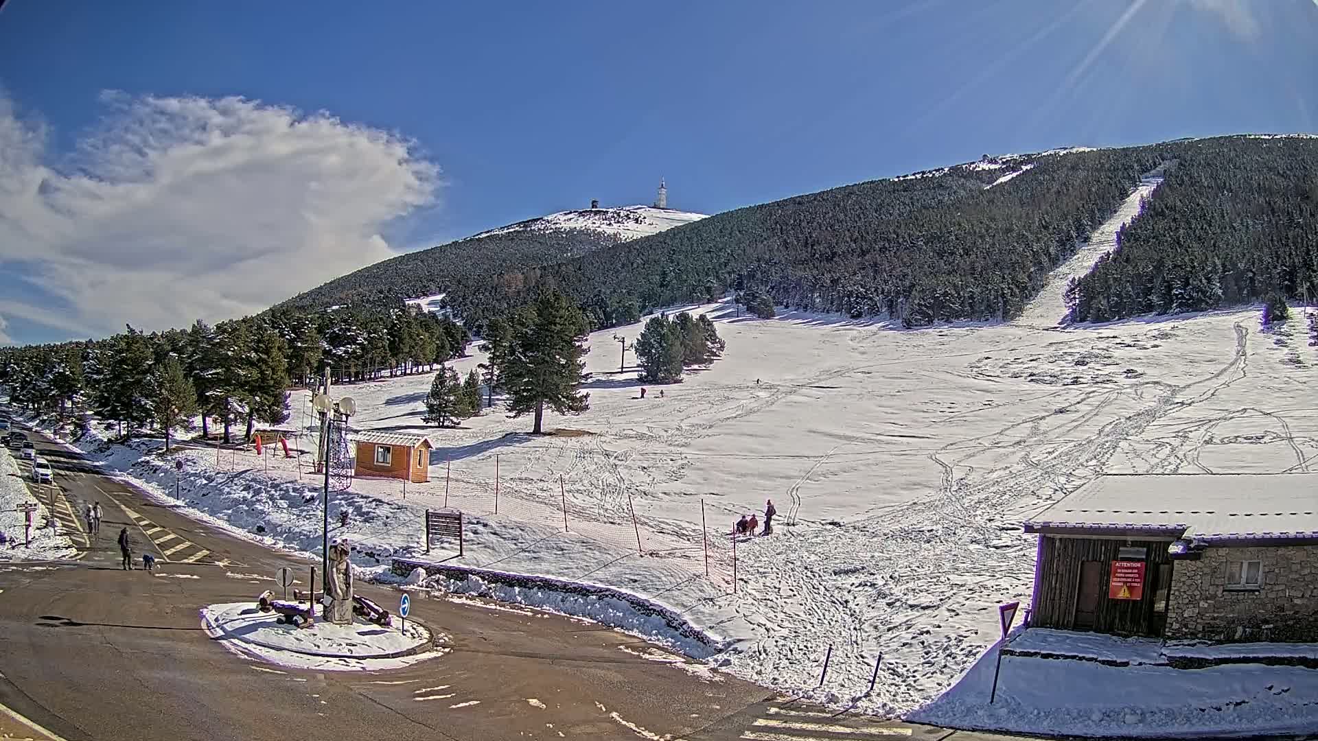 A snow-covered ski slope with several people on it is situated below a forested mountain on a sunny day with a few clouds.
