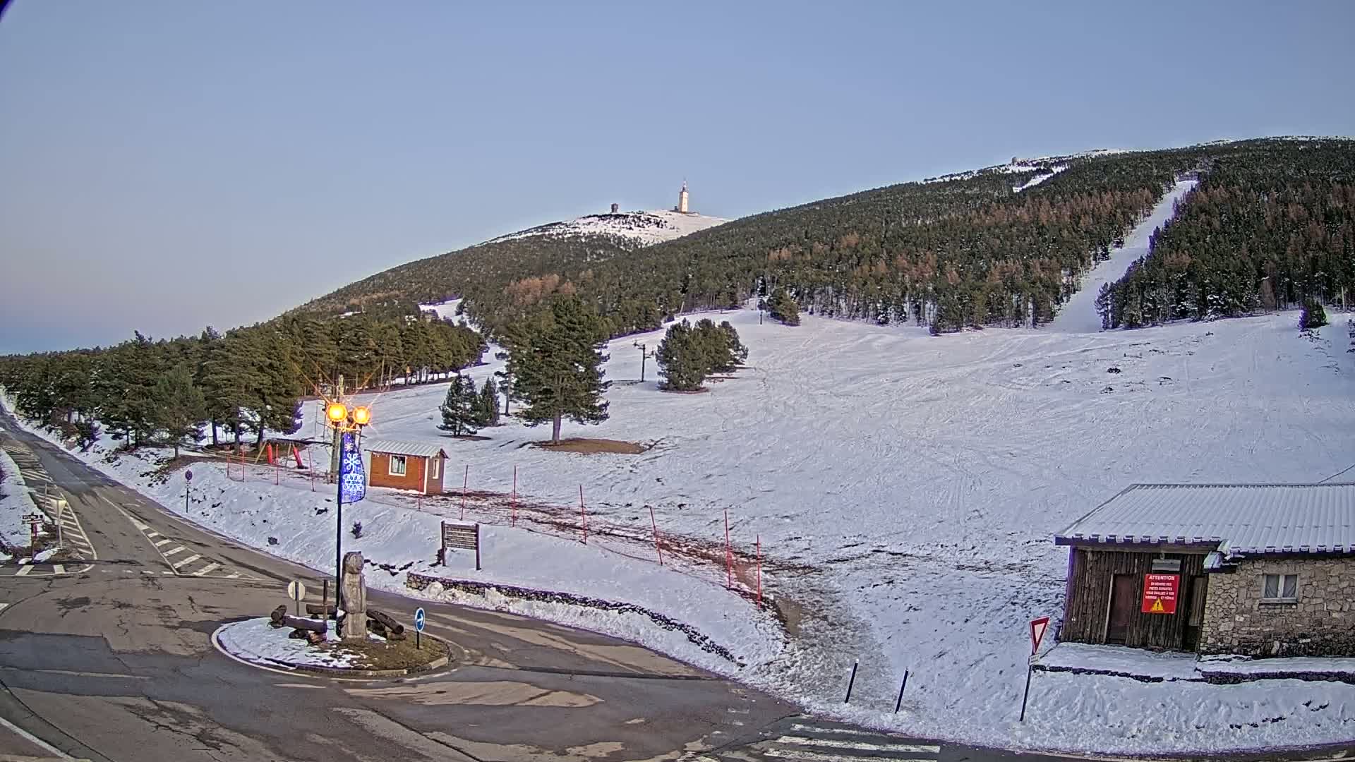 A snow-covered ski slope with a tall tower on a distant mountain peak under a clear, light-blue sky.