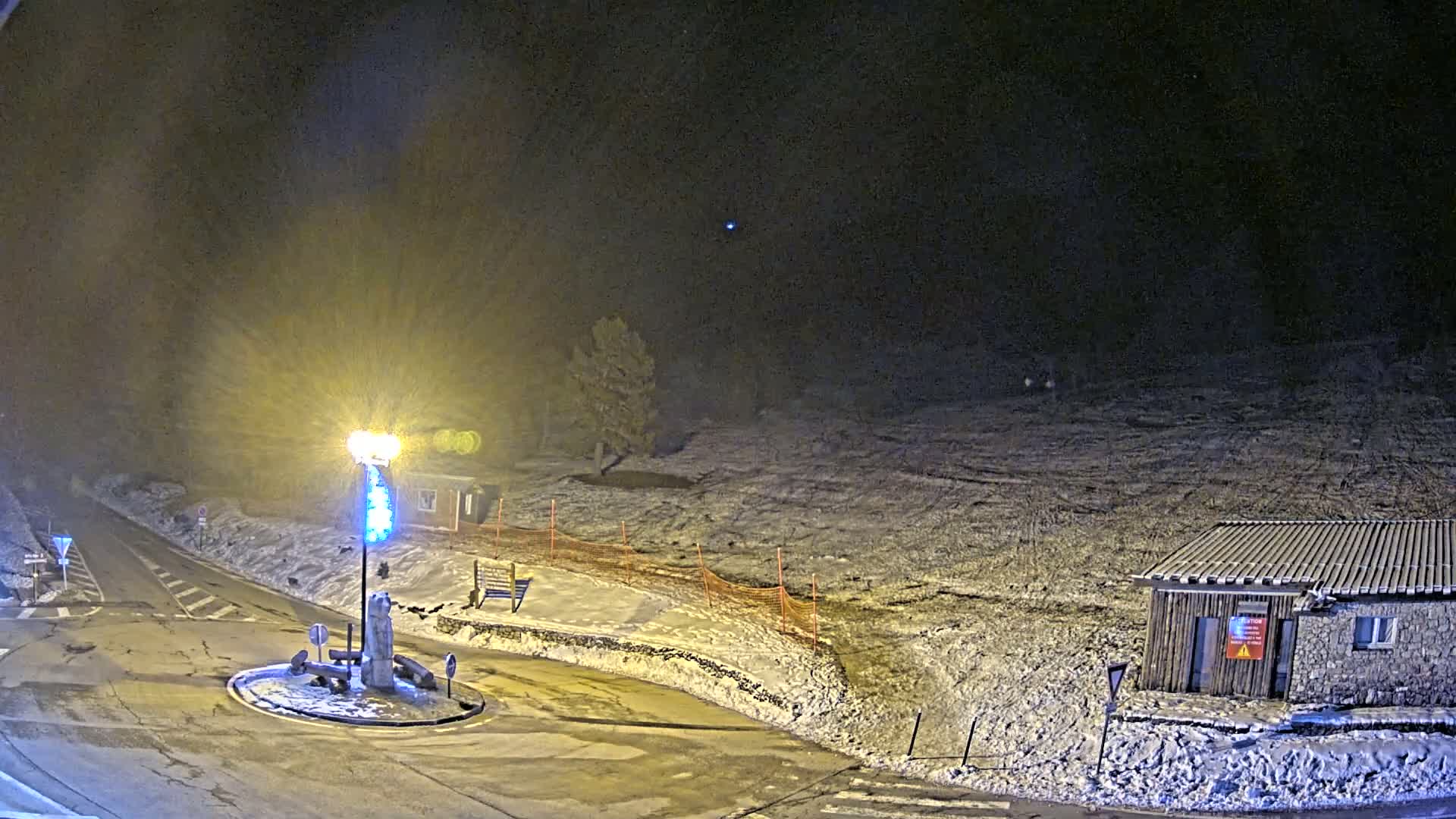 A snow-covered landscape at night is illuminated by a bright lamp at a roundabout near a small building.