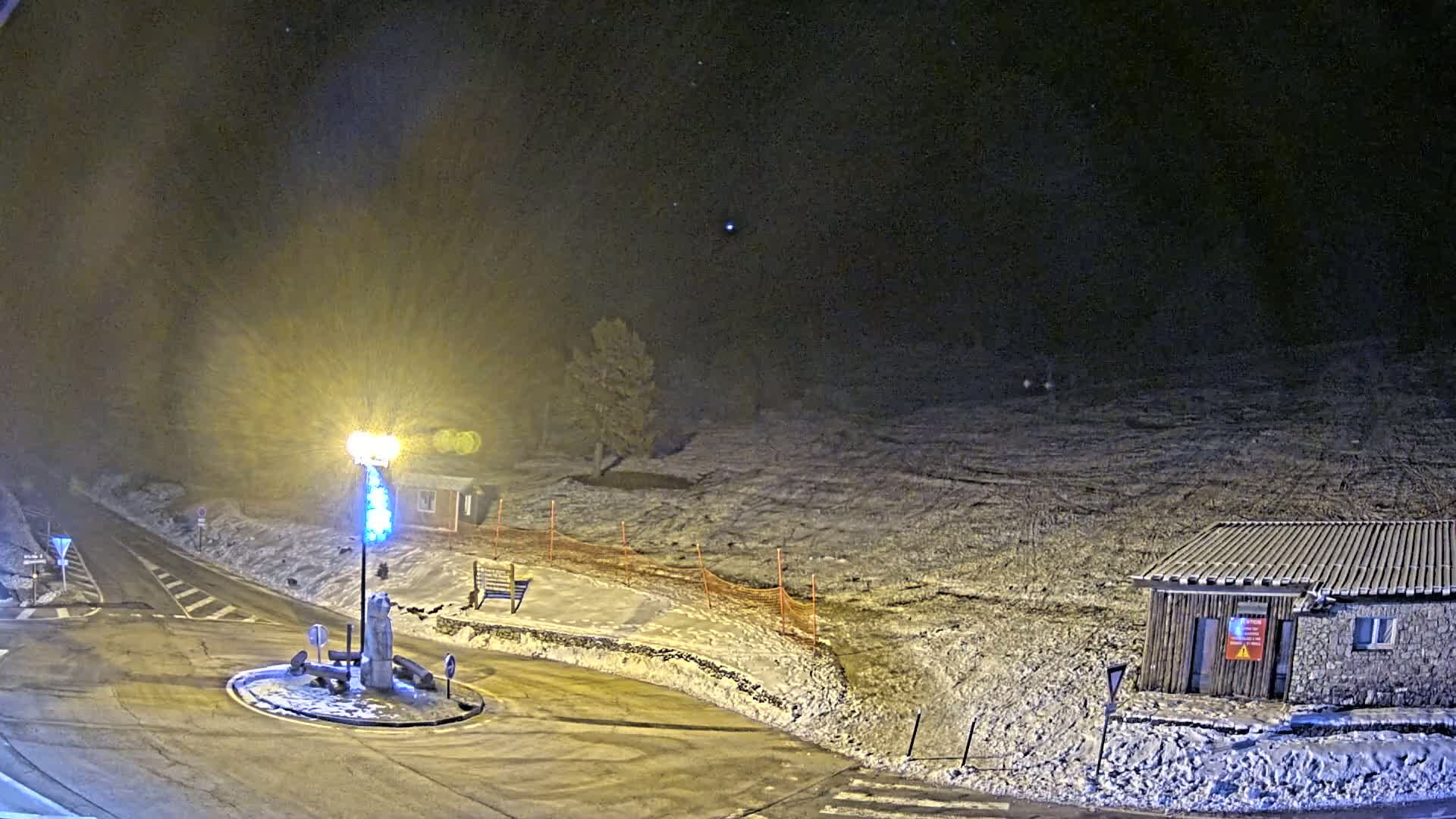 A snow-covered landscape at night is illuminated by a bright lamp at a roundabout near a small building.
