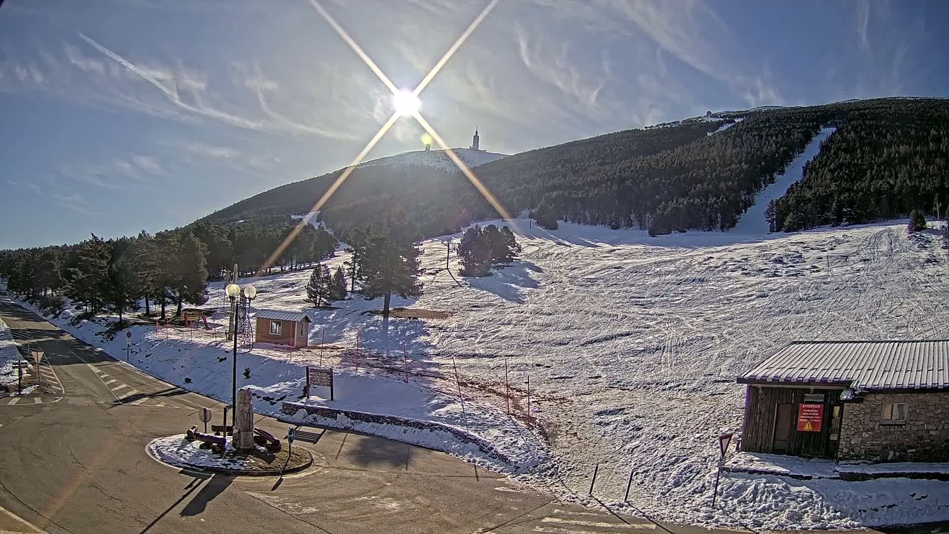 A snow-covered ski slope with a tall tower on a mountain in the background, under a sunny sky with some clouds.