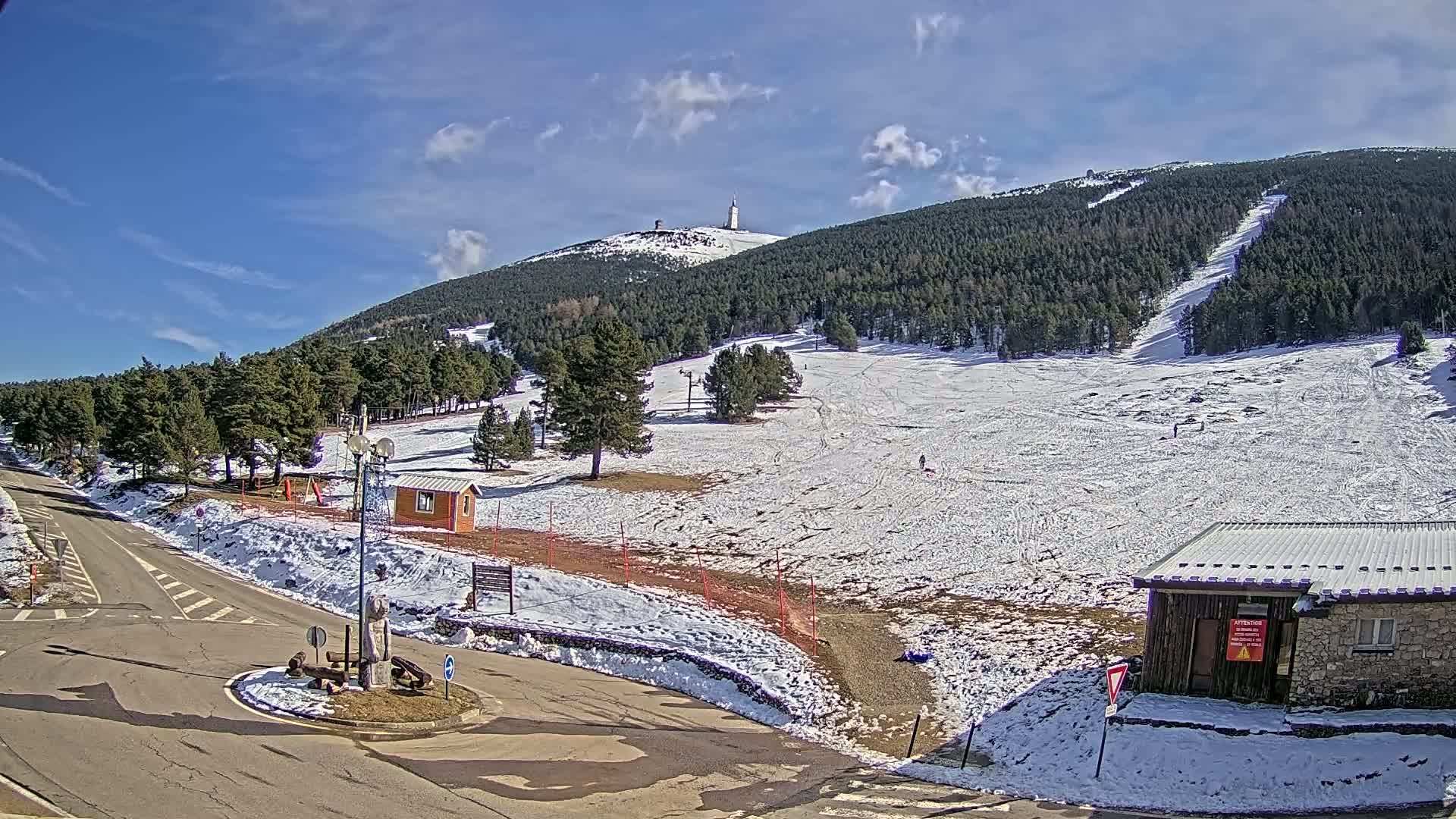 A snow-covered ski slope on a sunny day, with a small building and a road at the bottom, and a mountain with a tall tower in the background.