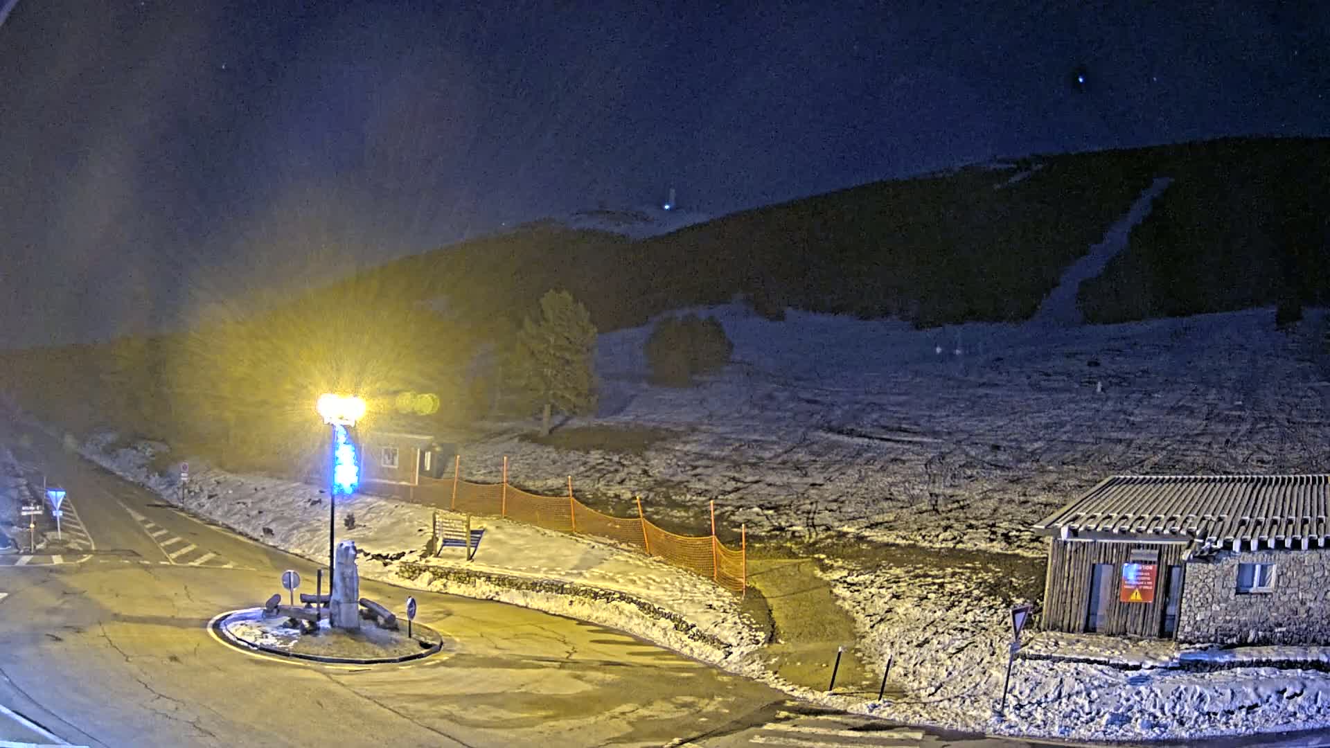 A snow-covered hill at night is illuminated by a bright lamppost near a small building and a roundabout.