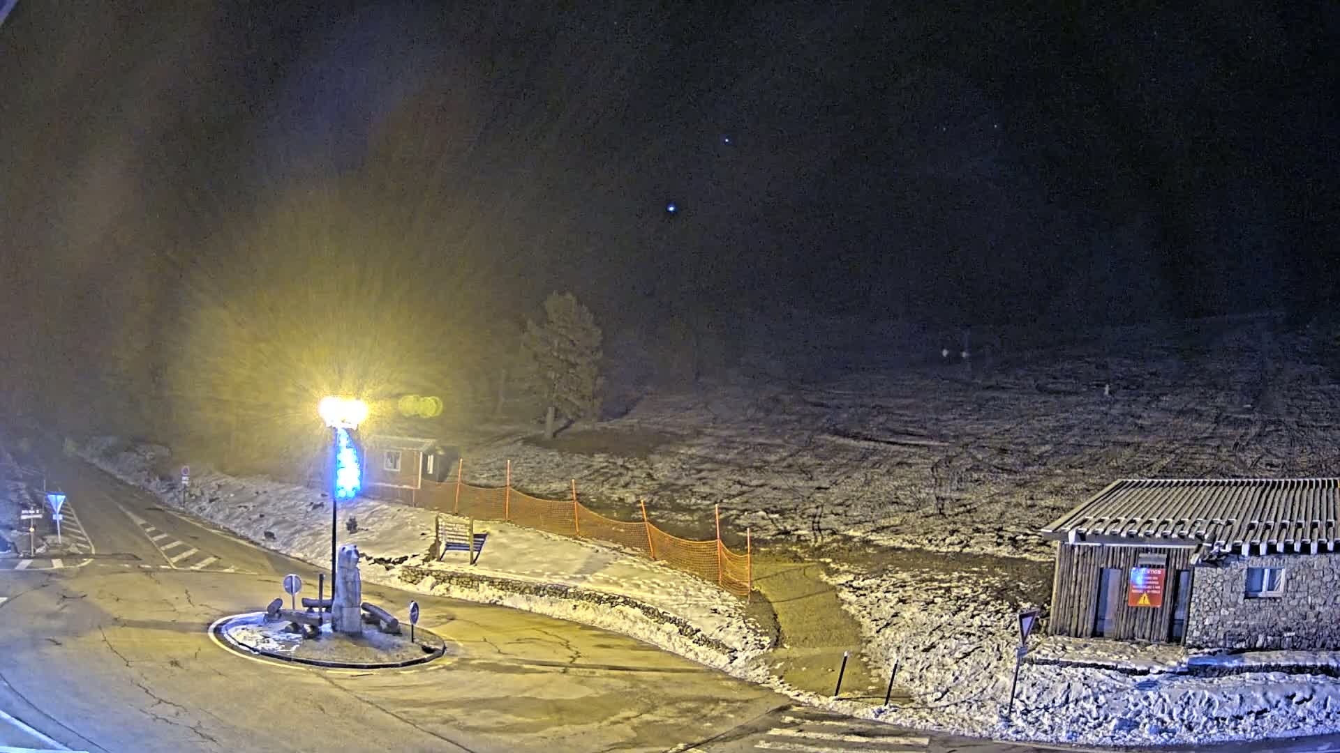 A snow-covered roundabout at night, illuminated by a bright lamppost, with a small building and orange safety netting visible in the background under a dark, mostly clear sky.