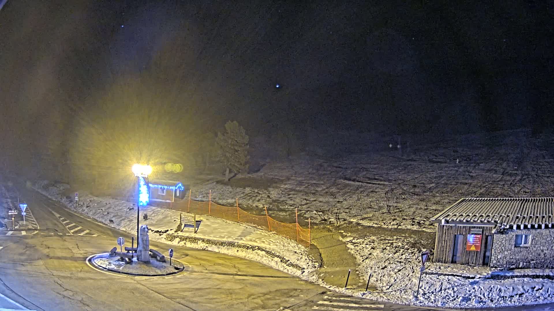 A snow-covered roundabout at night is illuminated by a bright lamppost, with a small building and a section of orange snow fencing visible in the background under a dark, clear sky.