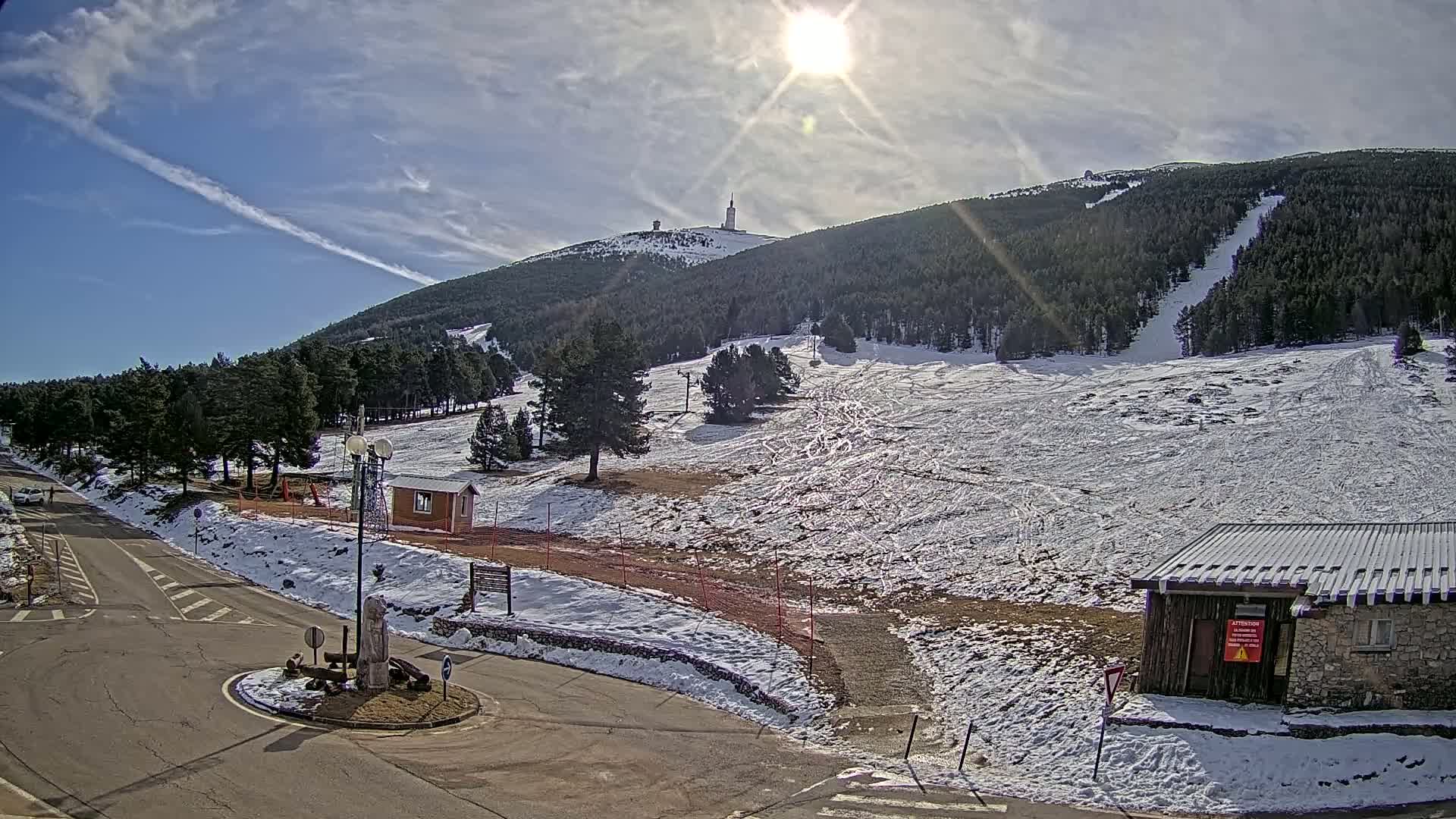 A sunny day at a snow-covered ski resort shows a mountain with a tall tower on its peak, along with a road, small buildings, and sparsely populated ski slopes.