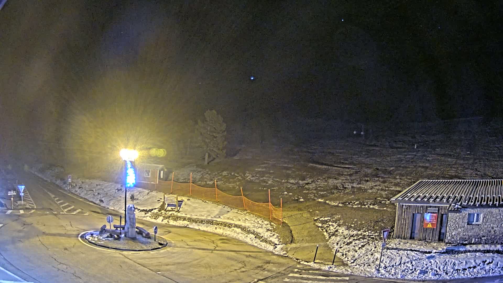 A nighttime, snow-dusted scene shows a roundabout and a small building near a sparsely vegetated area under a dark, clear sky.