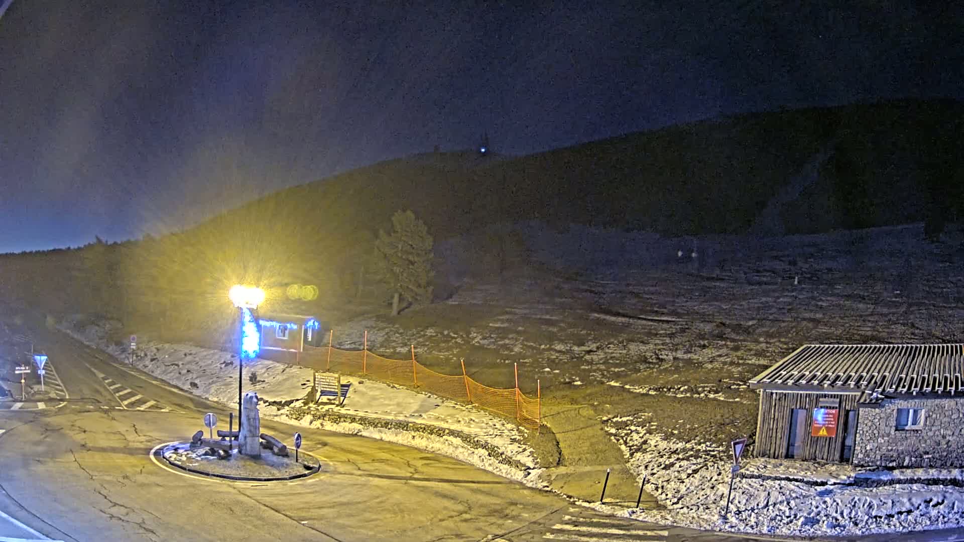 A nighttime scene shows a snow-dusted road intersection with a small building and a hill in the background under a dark sky.