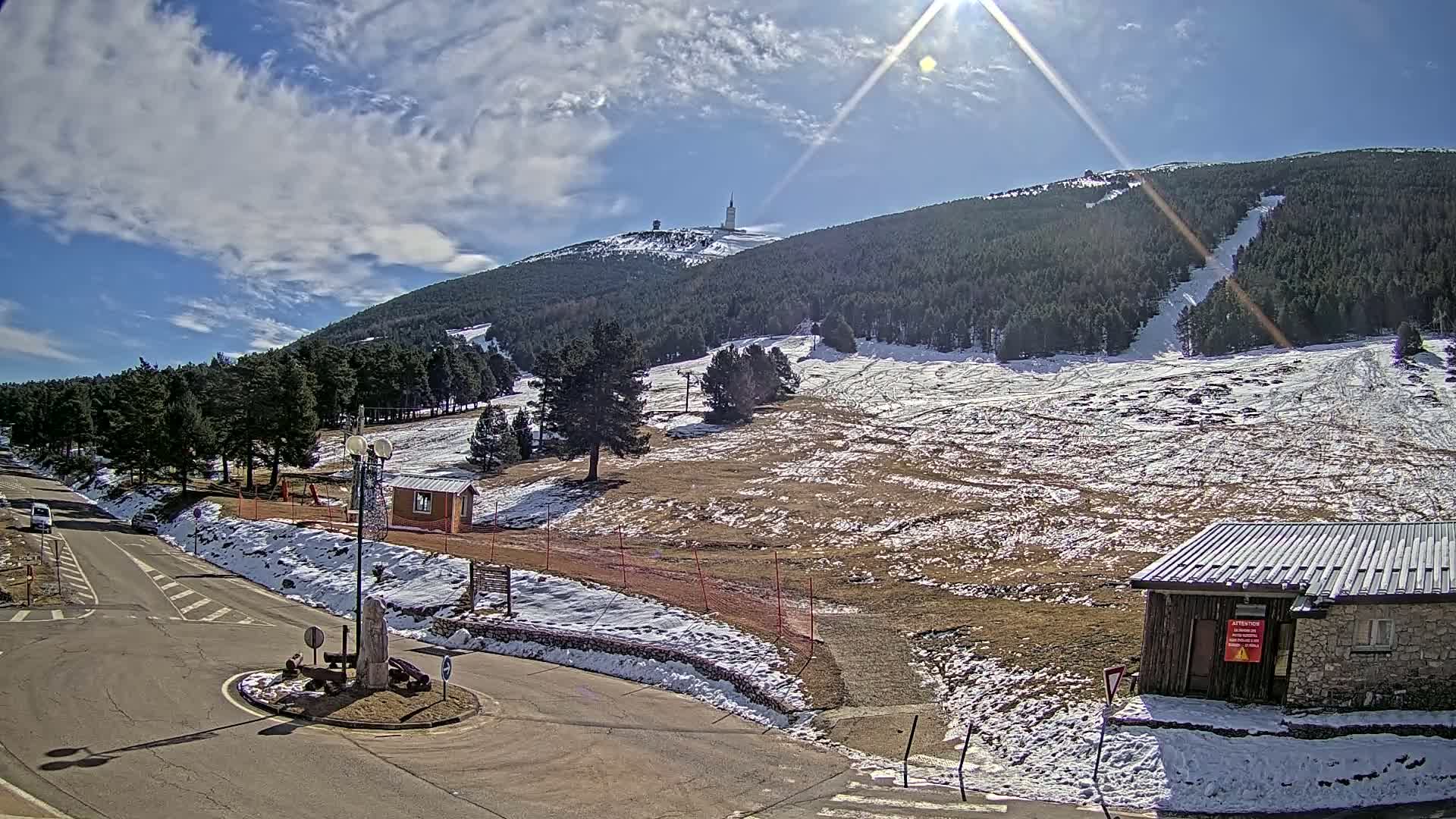 A sunny day with partly cloudy skies shows a snow-dusted mountainside with a tall tower at its peak, a small building, and roads below.