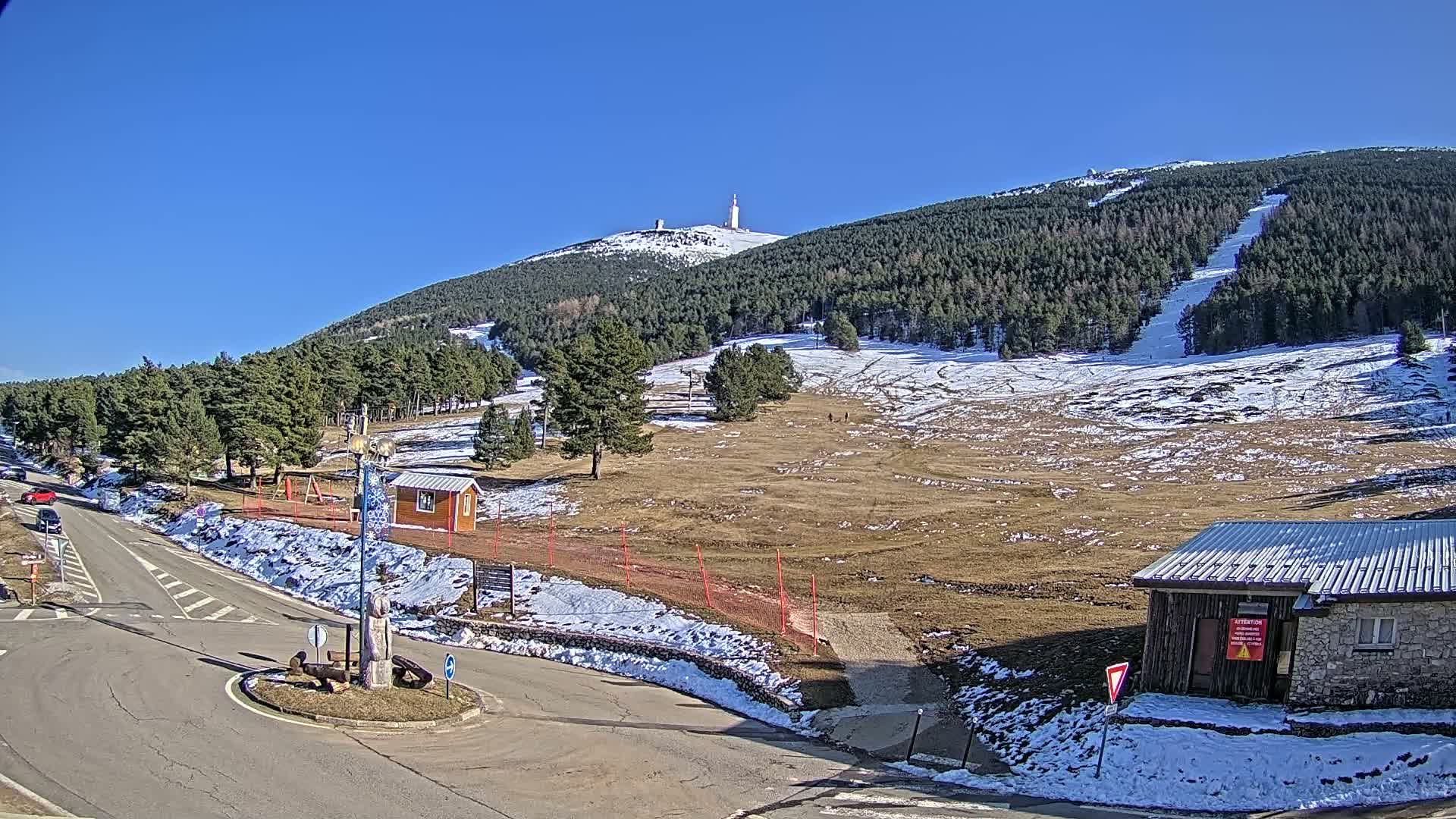A mountaintop with a communications tower is visible in the distance, beyond a partly snow-covered ski slope and a roadway, all under a clear blue sky.