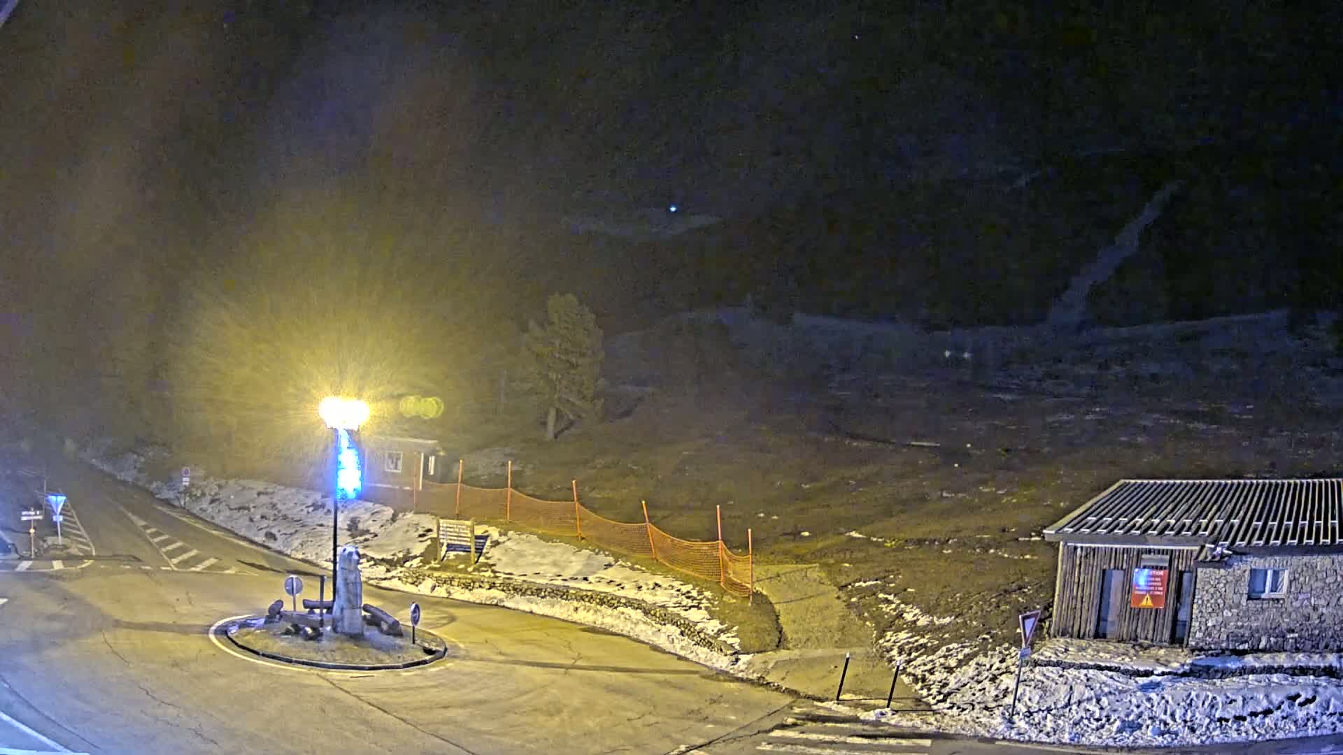 A nighttime view of a snow-dusted roundabout and a small building, illuminated by a bright lamppost, with a dark, hilly landscape in the background under a partly cloudy sky.