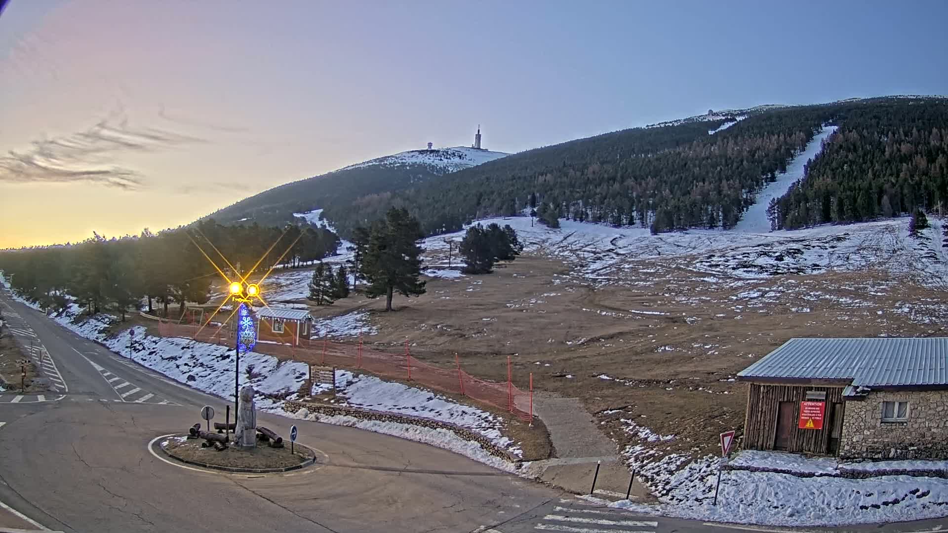A snow-dusted mountain slope with a tall tower atop, viewed from a road intersection at sunset.