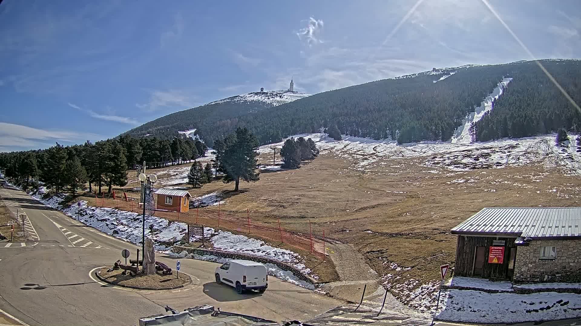 A sunny day at a mountain resort shows a mostly snow-covered ski slope with a tall tower atop the mountain in the background, while a van is parked near a small building in the foreground.