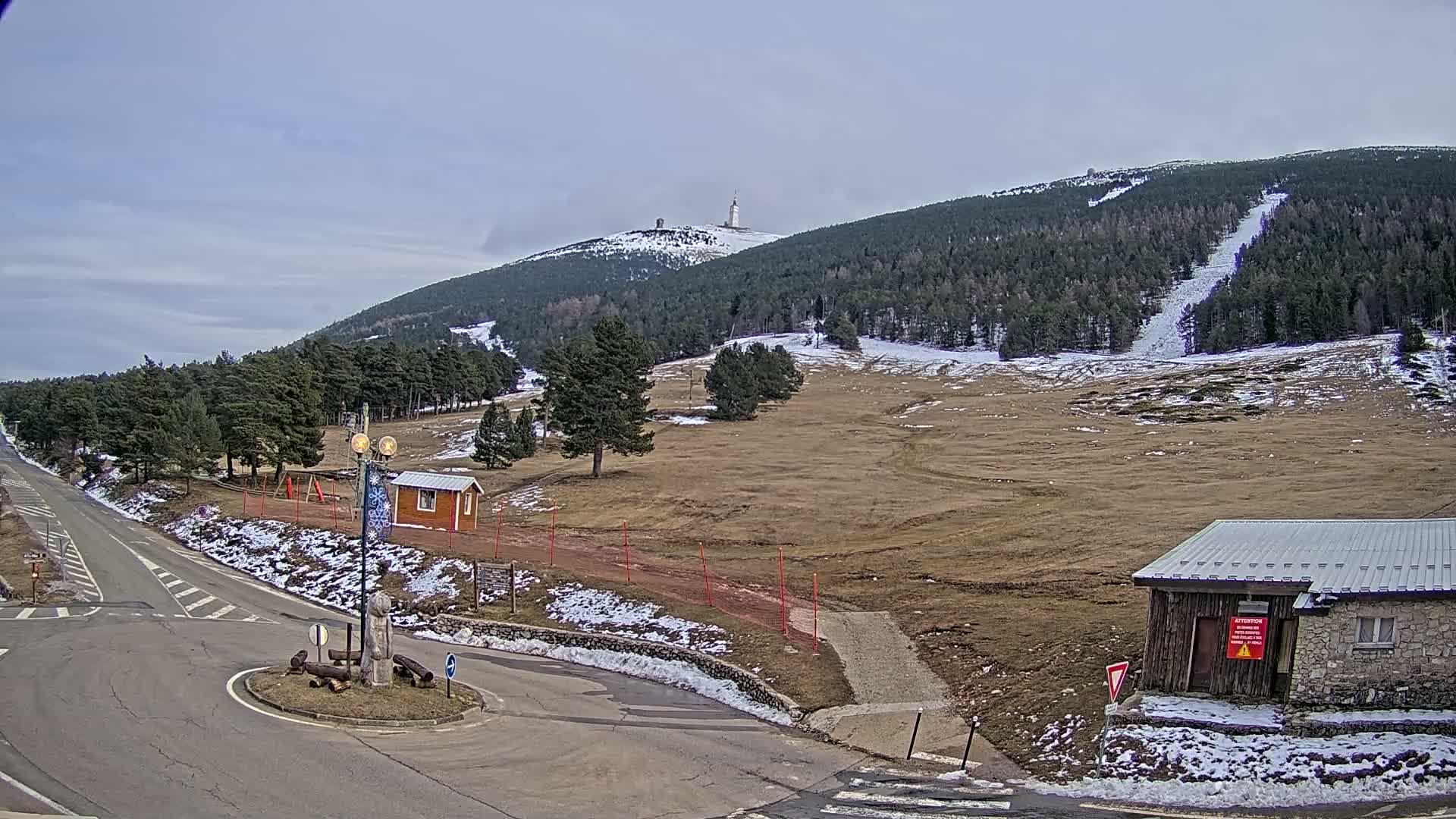 A mountaintop with a tall tower is visible in the distance, under a mostly cloudy sky, overlooking a sparsely snow-covered landscape with a road and small buildings near a roundabout.
